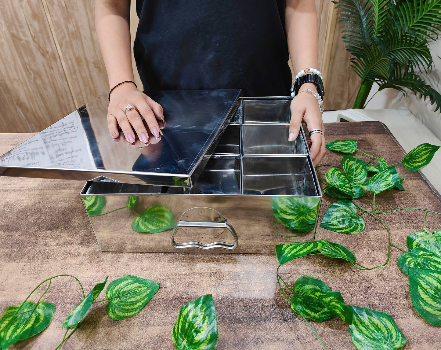 Person using a metal tray with compartments on a wooden surface with green leaves.