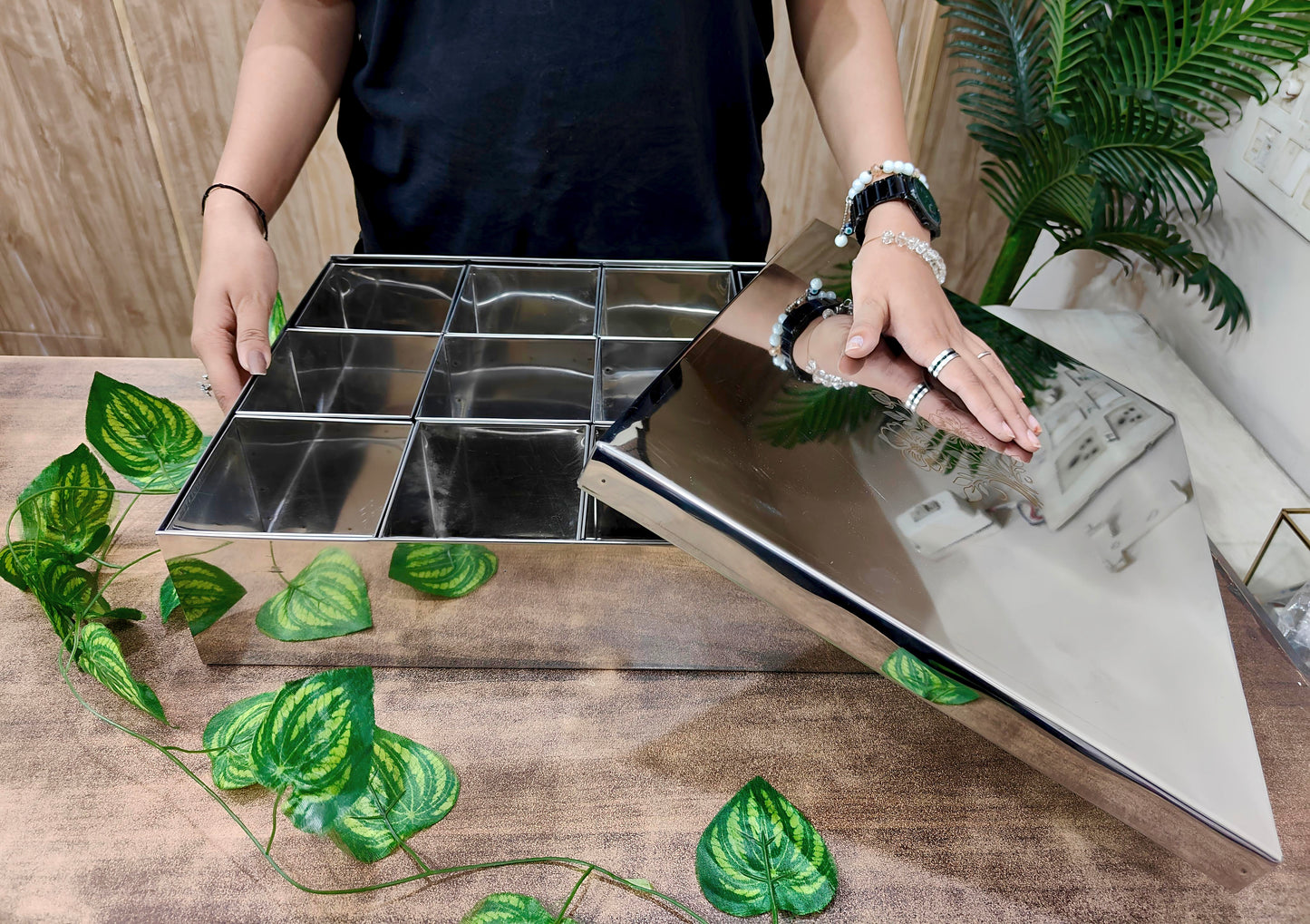 Person holding a metallic tray with compartments on a wooden surface with green leaves.