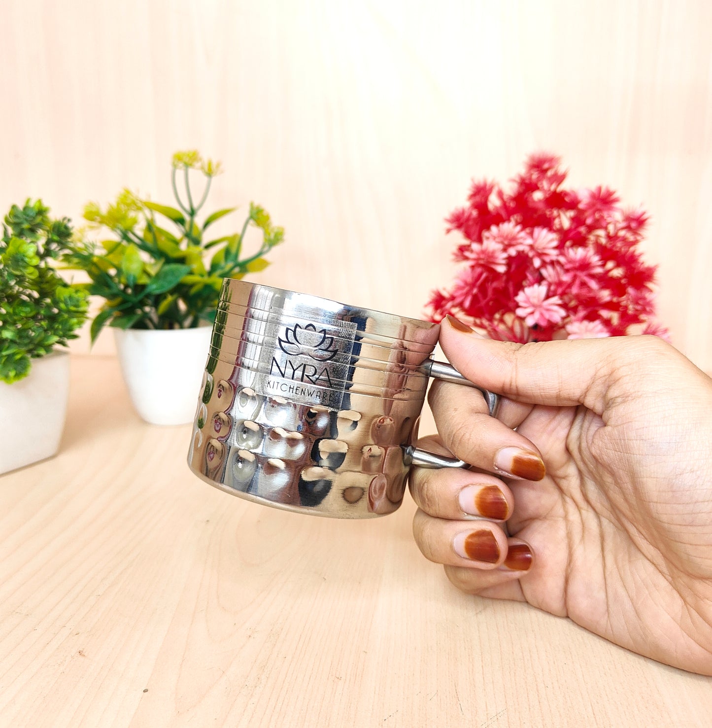Silver measuring cup with 'NYRA' branding held by a hand on a light surface with plants in the background