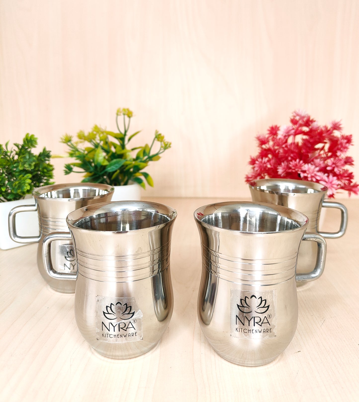 Set of silver mugs with 'Nyra' branding on a light background with plants.