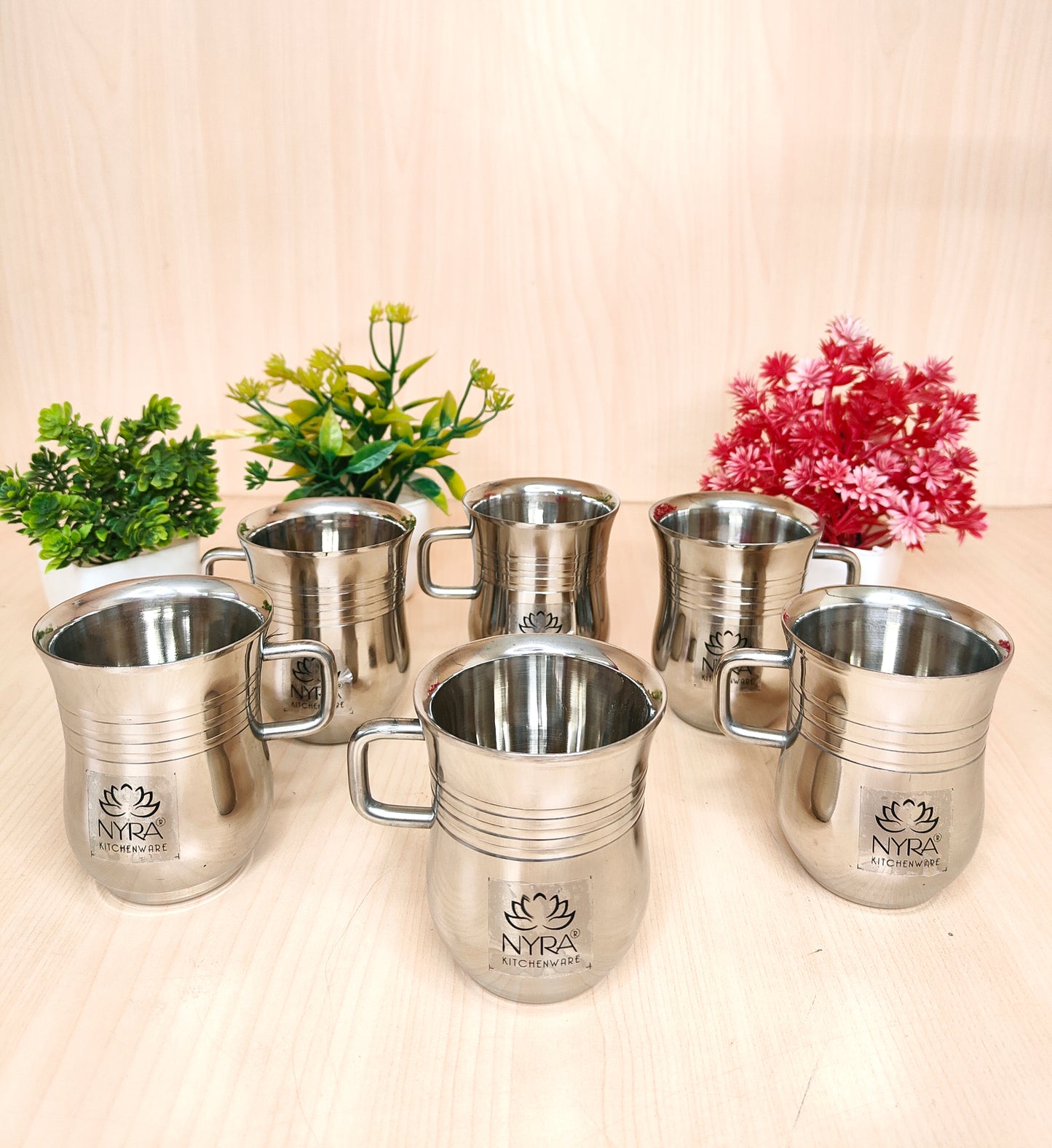 Set of silver mugs with 'NYRA' branding on a light wooden surface with plants.