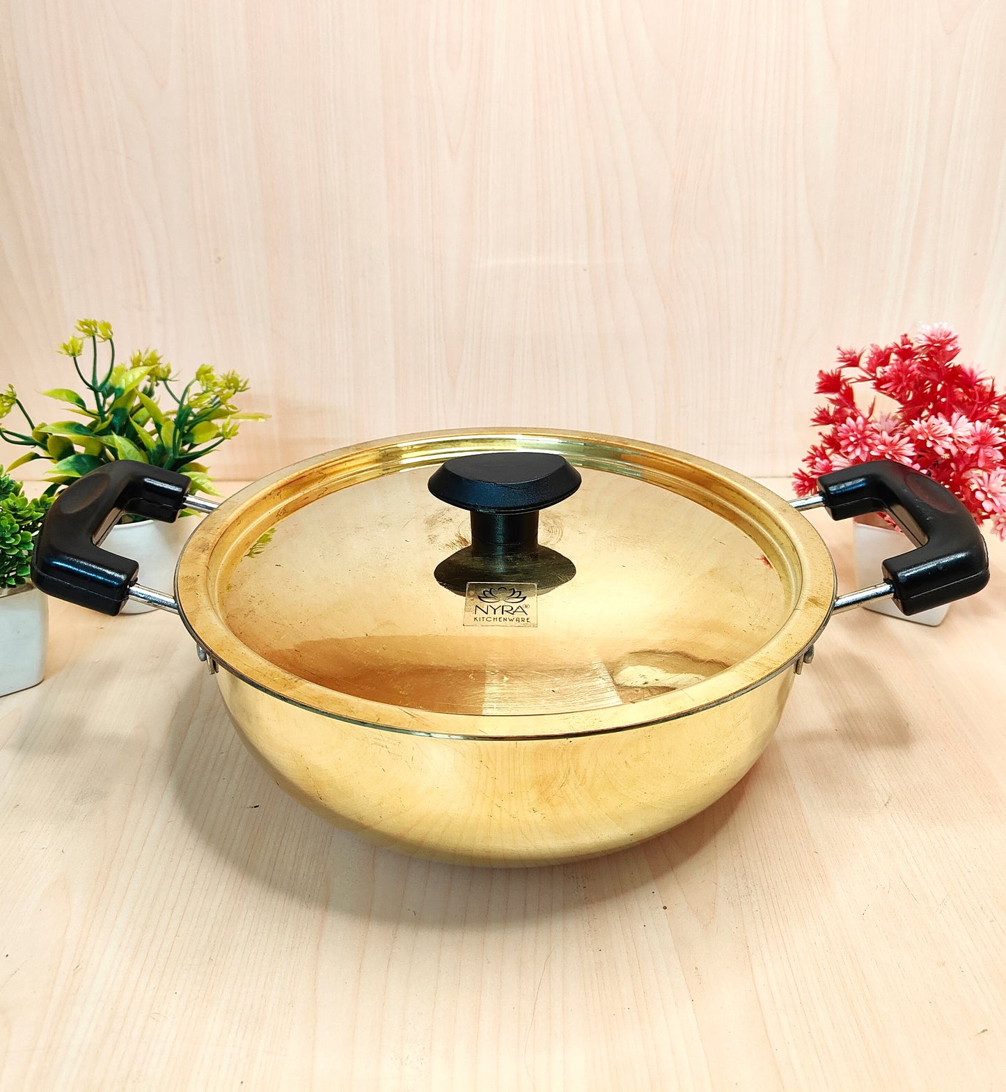Gold-colored pot with a lid on a light wooden surface with decorative plants.
