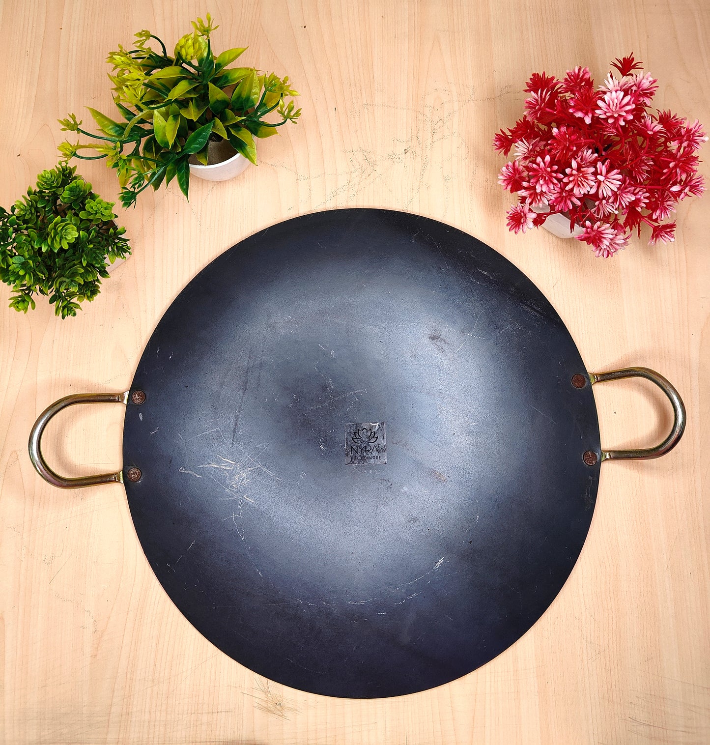 Black wok with handles on a wooden surface with decorative plants.