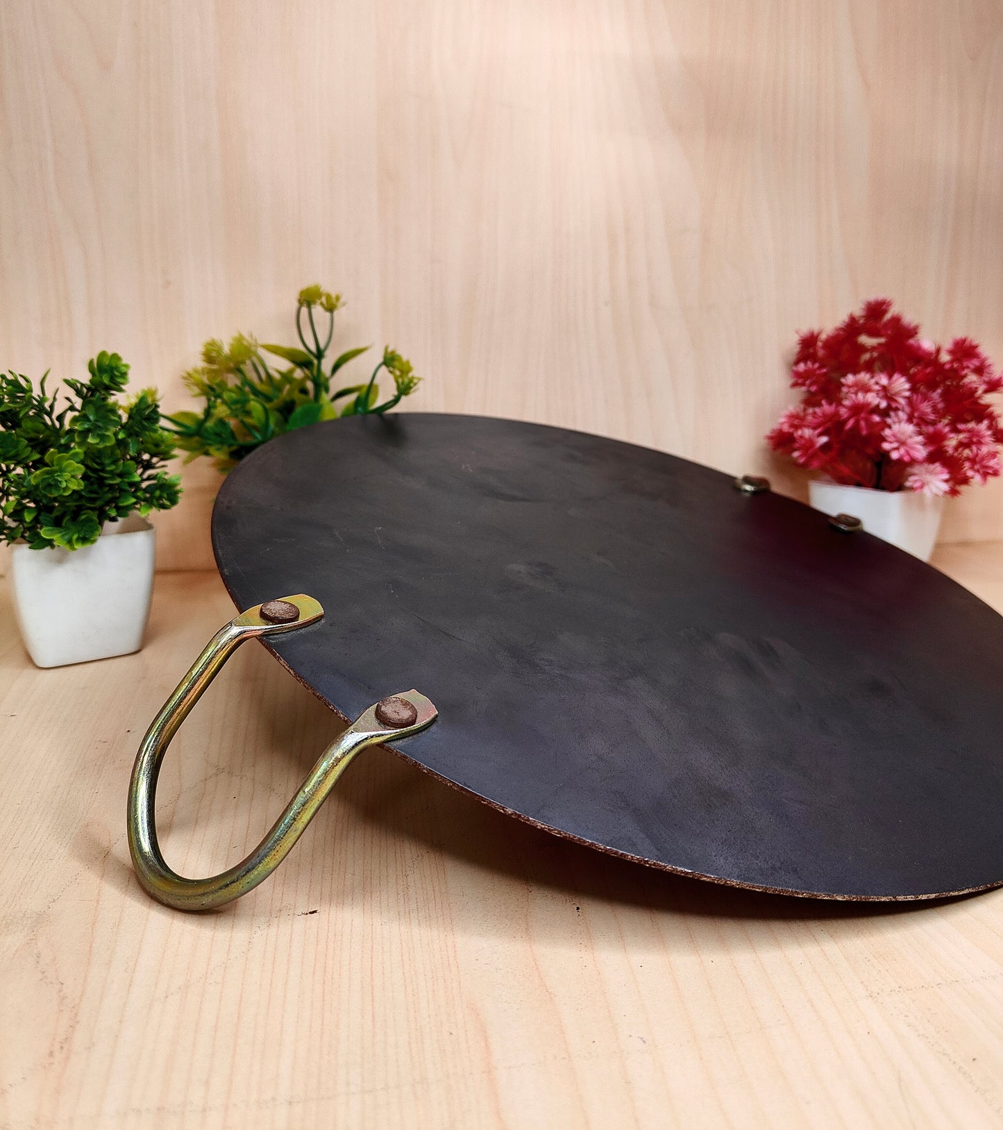 Black round fire pit with brass handle on a wooden surface with plants in the background
