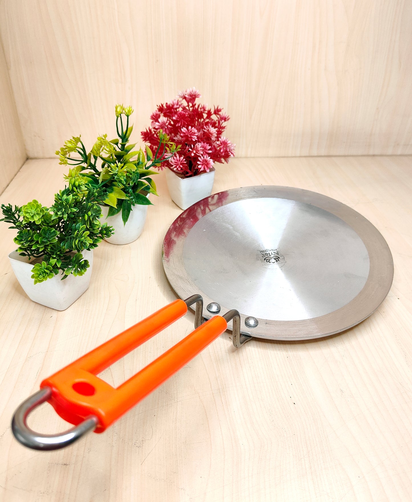 Metal camping pan with orange handle on a light wooden surface with plants in the background