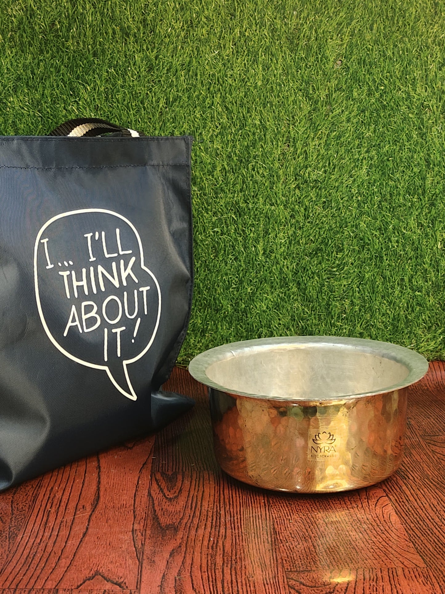 Black tote bag with 'I'll Think About It' text and a brass bowl on a wooden surface with grass background