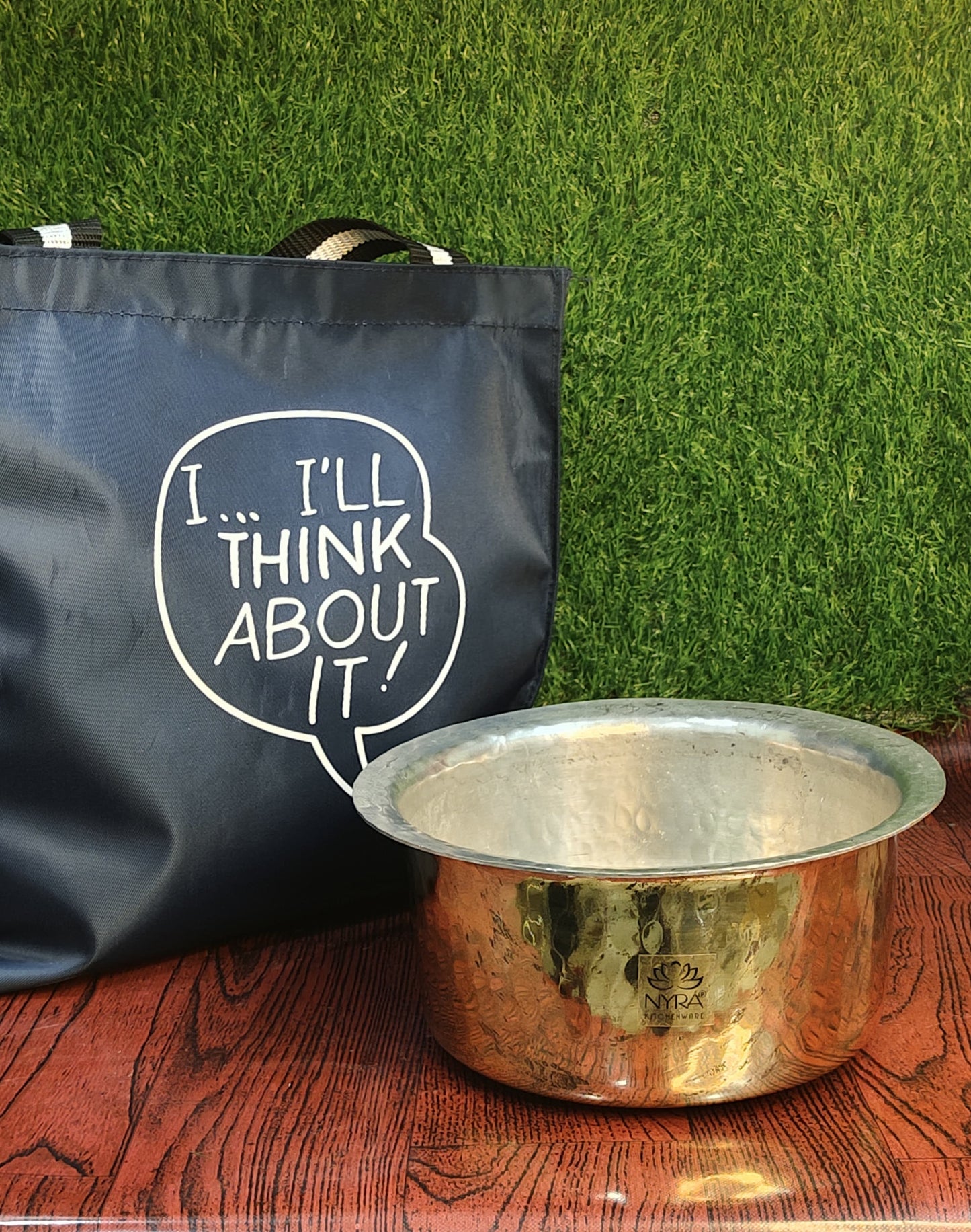 Copper pot and black bag with text on a wooden surface and green grass background
