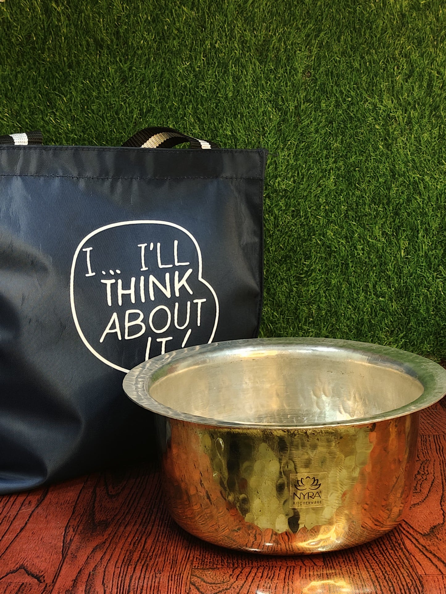 Black bag with text and a metallic bowl on a wooden surface against a green grass-like background