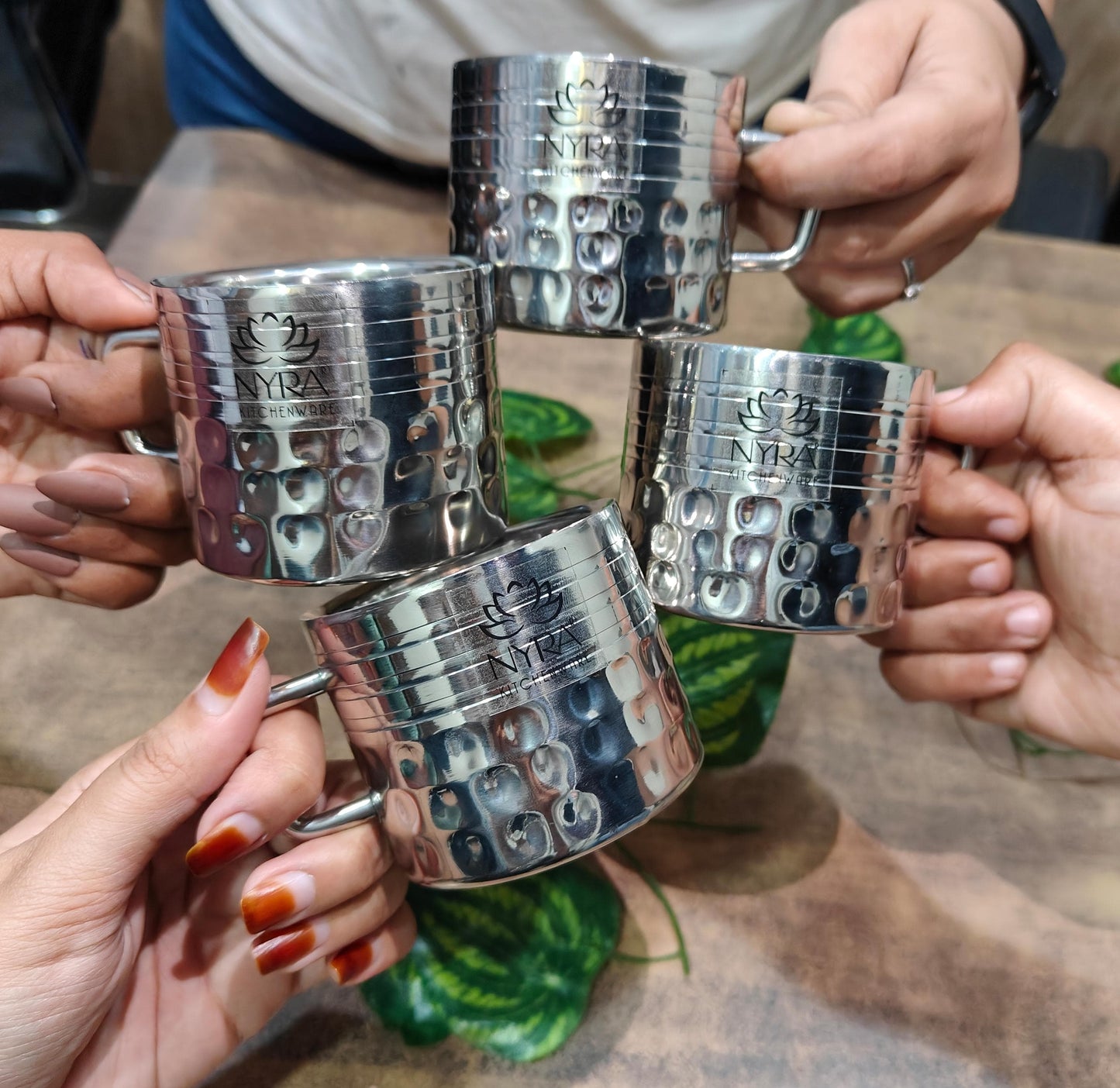 Four silver mugs with a textured surface held by hands on a wooden table.