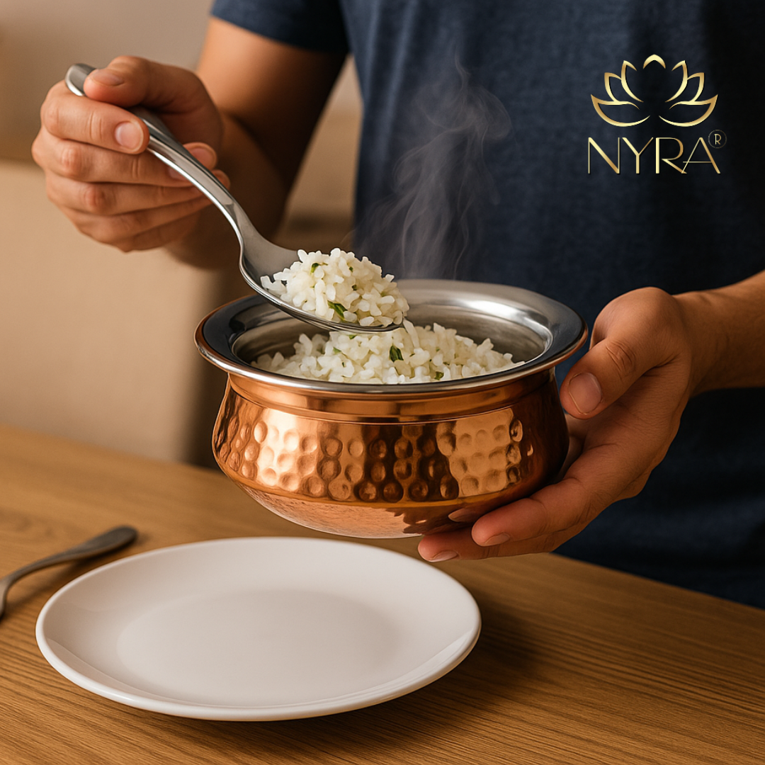 Person holding a copper bowl with steaming rice, using a spoon, on a wooden table with 'NYRA' branding.