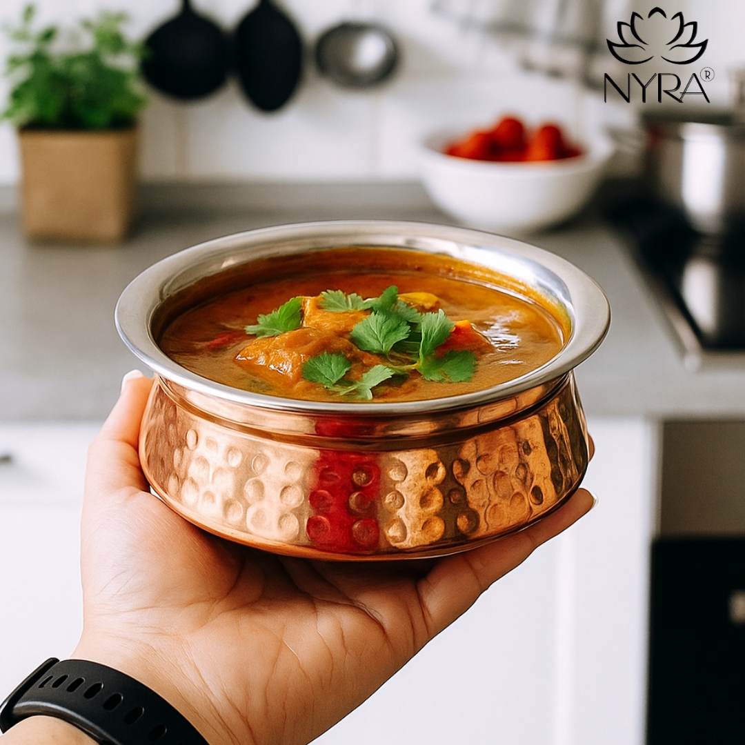Copper bowl of curry held by a hand with 'NYRA' brand logo in the background
