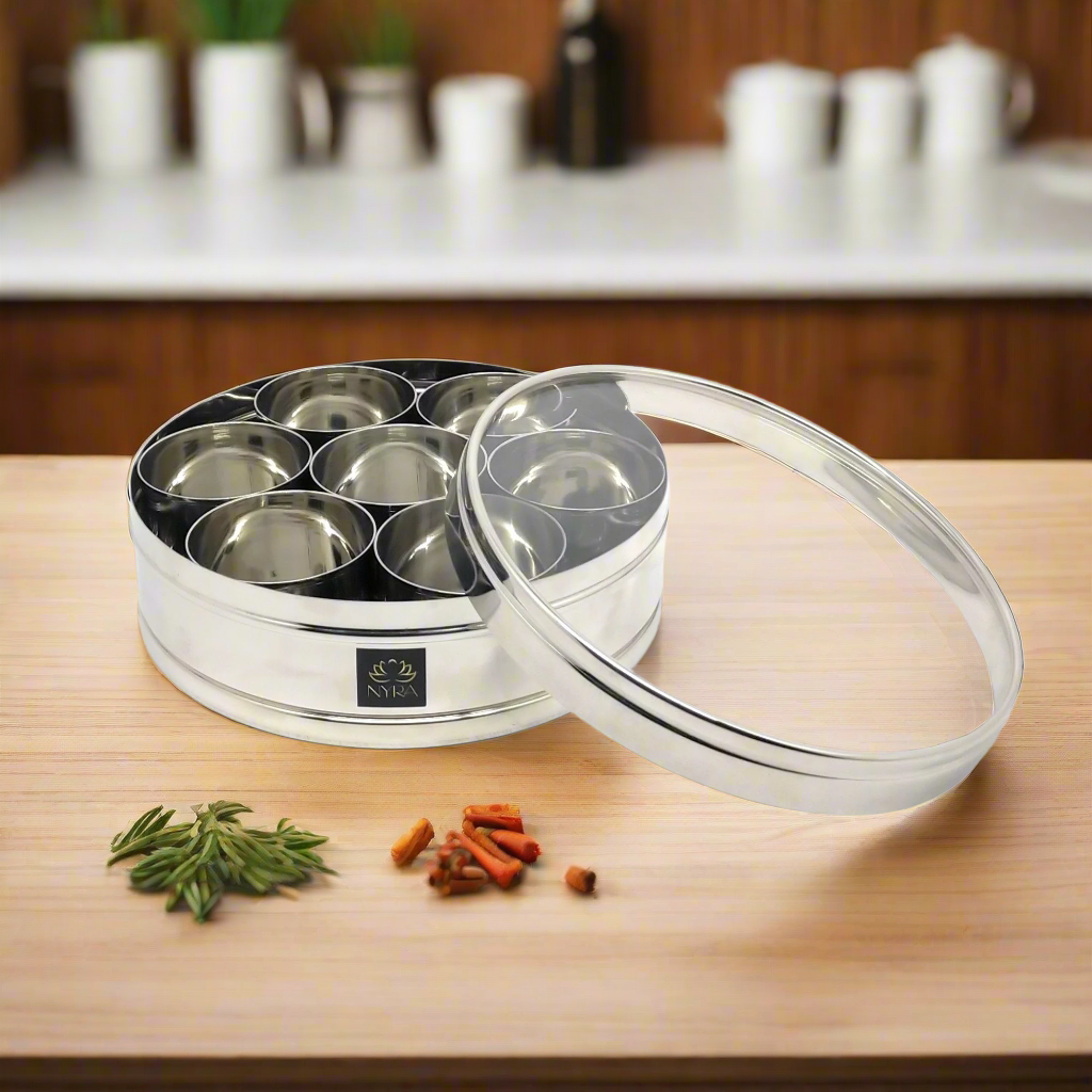 A stainless steel spice box with a see-through lid and seven storing bowls, placed on a wooden surface with a few dried spices in front of it.