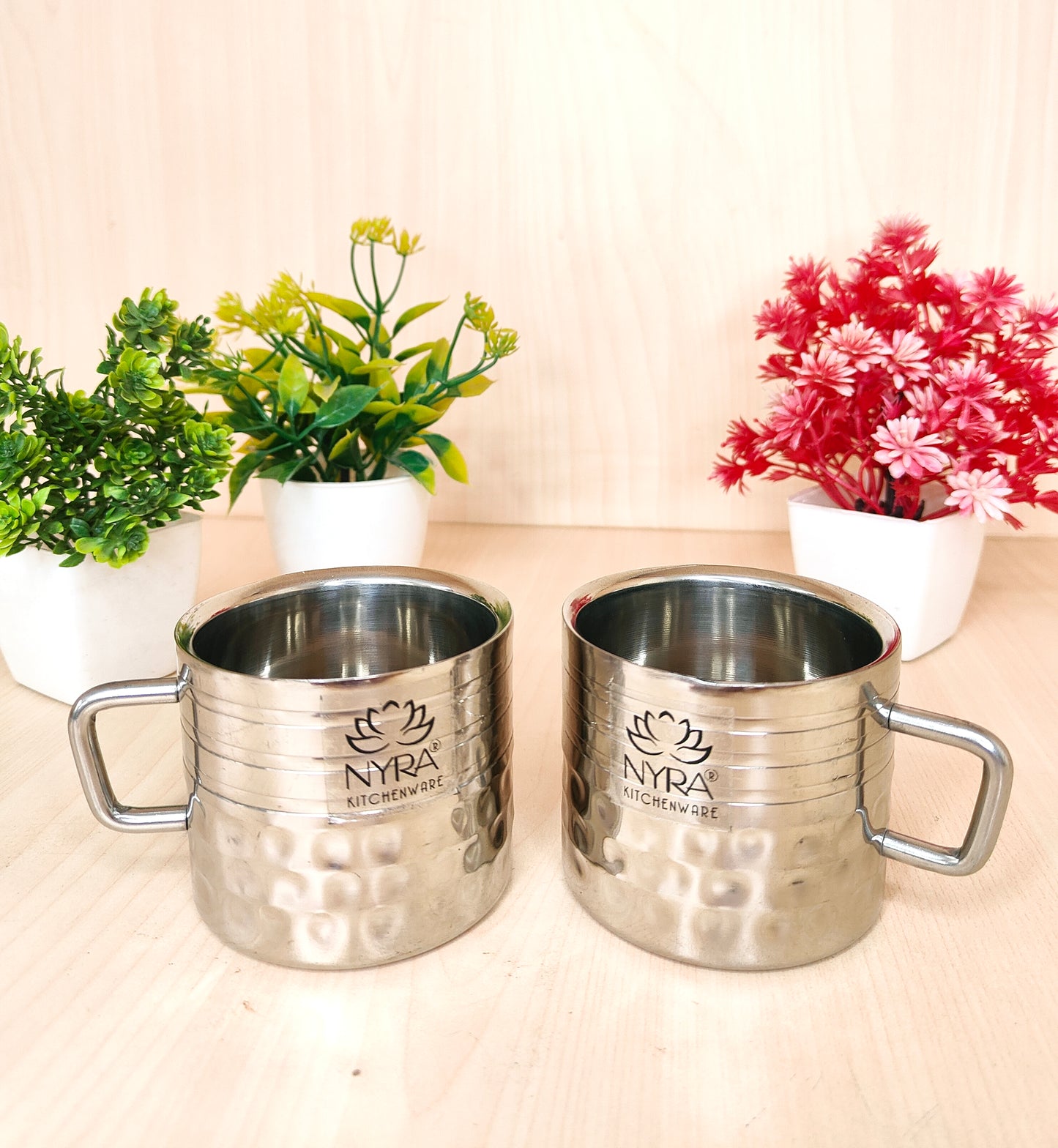 Two silver mugs with 'Nyra' branding on a light surface with plants in the background.