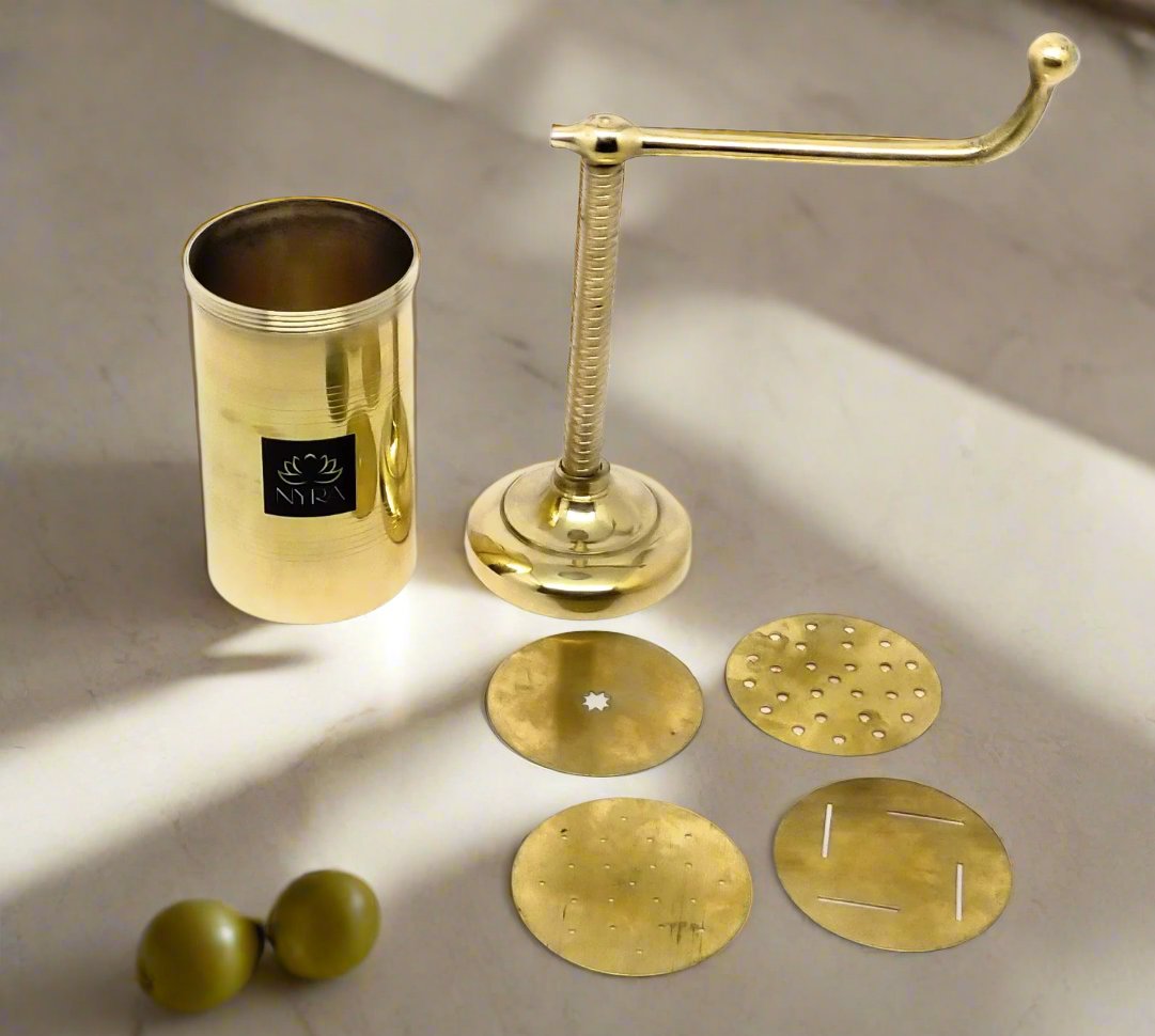 A brass olive press and mill set displayed on a light marble surface with dramatic shadows. The set includes a cylindrical press head with a black brand label, a curved press stem and anvil, four round brass plates of varying perforation patterns, and two olive pits.