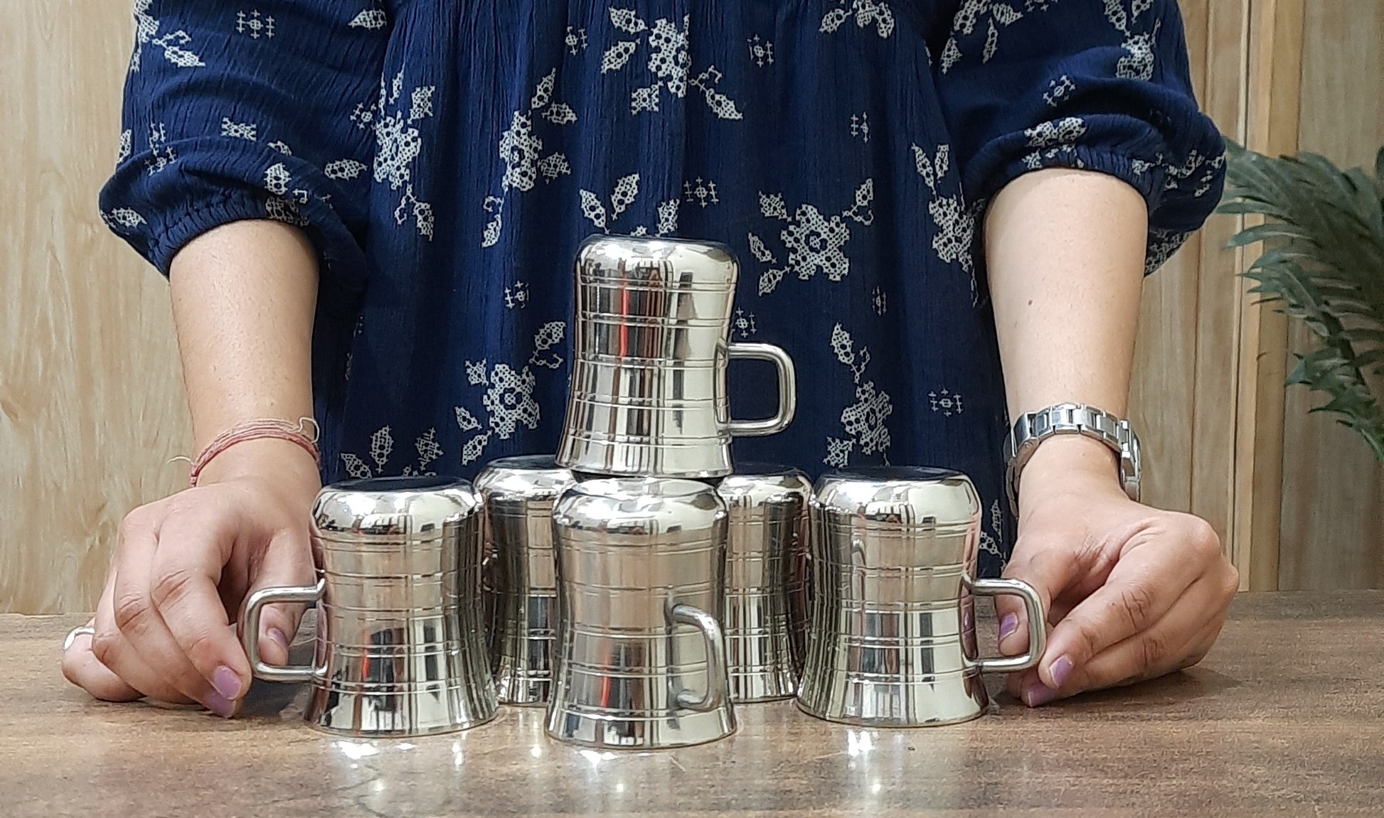 Two stainless steel double-walled mugs with a glossy finish are placed on a wooden surface. One mug is standing upright showing the exterior side, while the other mug is on its side, displaying the interior, which is silver-colored.