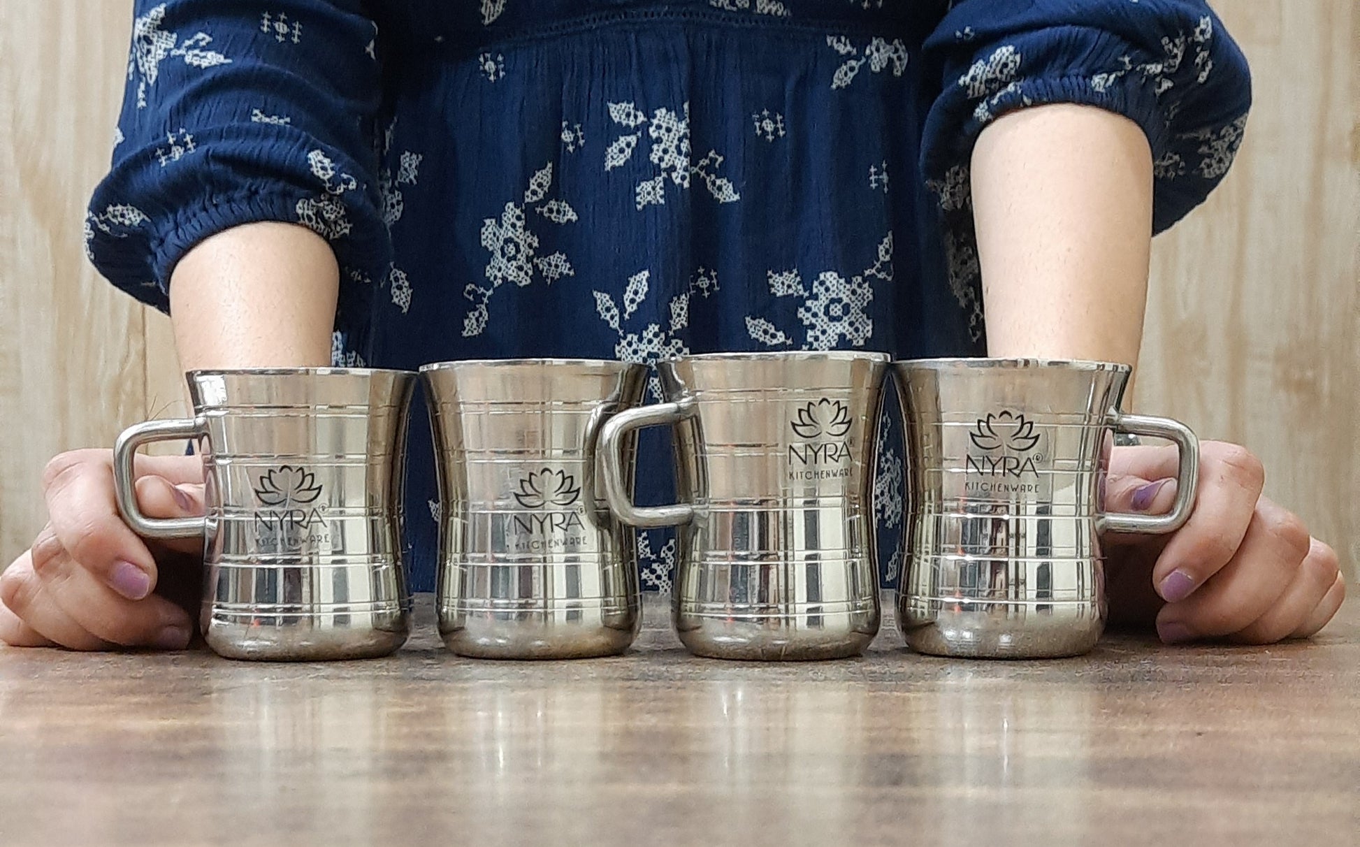 Two stainless steel double-walled mugs with a glossy finish are placed on a wooden surface. One mug is standing upright showing the exterior side, while the other mug is on its side, displaying the interior, which is silver-colored.