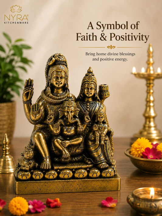 A bronze-toned multi-armed Hindu deity statue displayed on a wooden surface with lit oil lamps, flowers, and a warm neutral background. Text at the top reads 'A Symbol of Faith & Positivity' with the NYRA Kitchenware logo in the upper left.