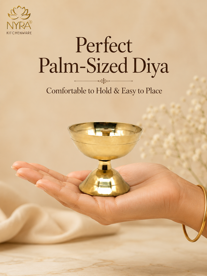 A brass diya holder being held in a person's hand, with text reading 'Perfect Palm-Sized Diya' and 'Comfortable to Hold & Easy to Place' on a warm beige background. The NYRA KITCHENWARE logo appears in the top left.