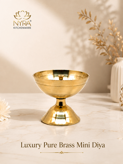 A polished gold brass mini diya holder with a pedestal base, displayed on a marble surface with warm decorative shadows and dried botanicals in the background. The NYRA Kitchenware logo is visible in the top left, and product text at the bottom reads 'Luxury Pure Brass Mini Diya'.