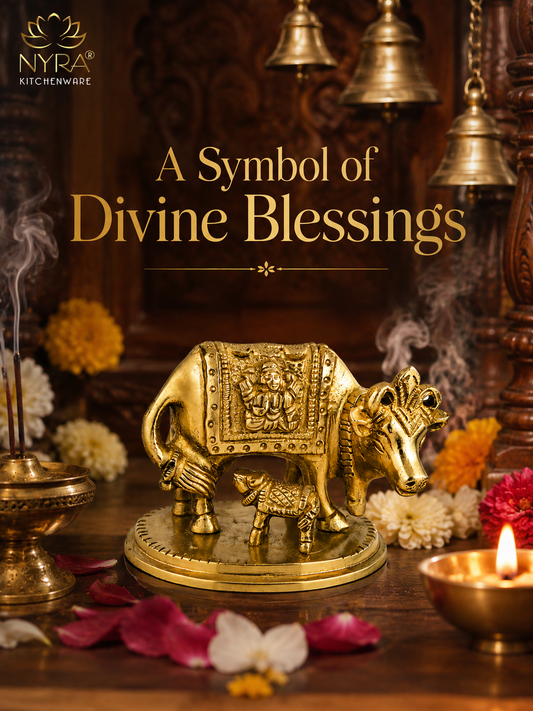 A brass cow and lamb figurine on a round base, displayed on a wooden altar with marigold flowers, a burning brass oil lamp, incense, and hanging brass bells in the background. The NYRA Kitchenware logo is visible in the top left, and the text 'A Symbol of Divine Blessings' is overlaid in the center.