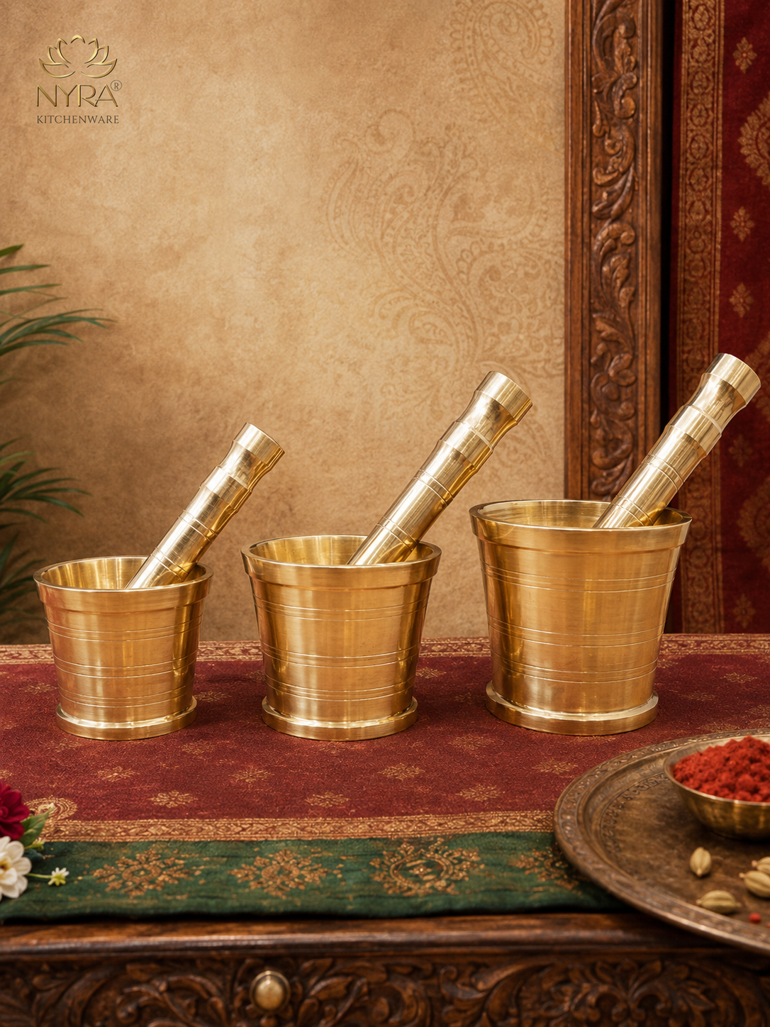 Three brass mortar and pestle sets arranged on a red patterned runner on a carved wooden table, with a decorative wall backdrop and a plate of red spices in the foreground. The brand logo 'NYRA KITCHENWARE' is visible in the top left corner.