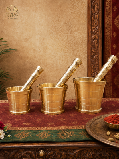 Three brass mortar and pestle sets arranged on a red patterned runner on a carved wooden table, with a decorative wall backdrop and a plate of red spices in the foreground. The brand logo 'NYRA KITCHENWARE' is visible in the top left corner.