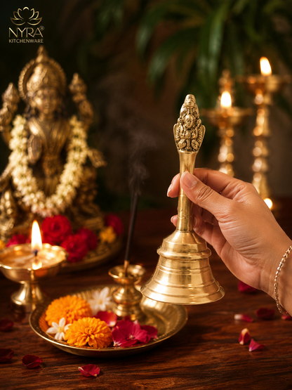 A hand holds a polished brass temple bell in a ceremonial setting with lit oil lamps, flowers, incense, and a decorative statue in the background. The NYRA Kitchenware logo is visible in the top left corner.