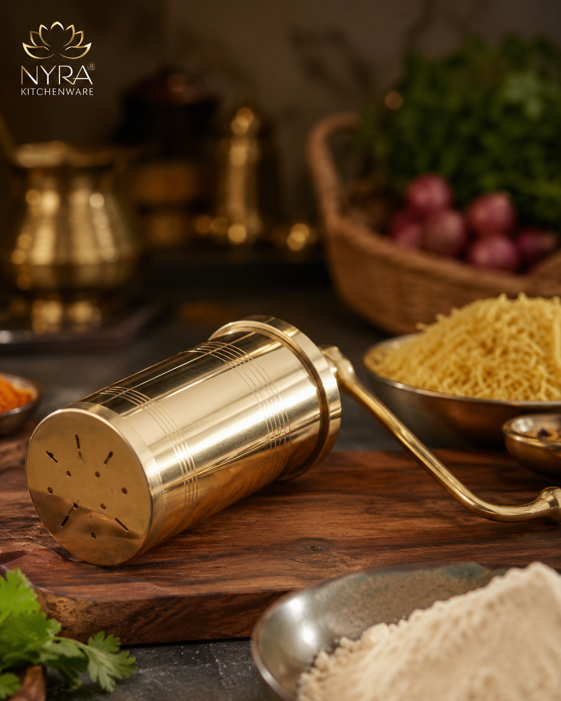 A brass spice grinder with a cylindrical body and perforated top, resting on a wooden board with bowls of ingredients and brass kitchenware in the background. The NYRA Kitchenware logo is visible in the top left corner.