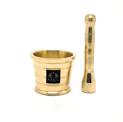 A gold-colored brass mortar and pestle set on a white background, featuring a black square logo plate on the mortar with the brand name 'NYRA'.