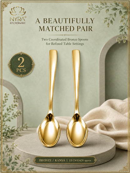 Two polished gold-toned bronze kitchen spoons displayed side by side on a round pedestal, with decorative text reading 'A BEAUTIFULLY MATCHED PAIR' and 'Two Coordinated Bronze Spoons for Refined Table Settings.' The NYRA Kitchenware logo appears in the top left, and the spoons are shown against a softly styled background with draped fabric and botanical accents.