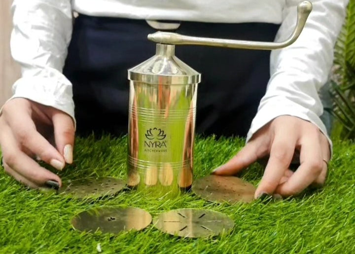 A person using a NYRA hand-crank lawn aerator on green grass, with the metallic gold cylinder and brass blades visible.