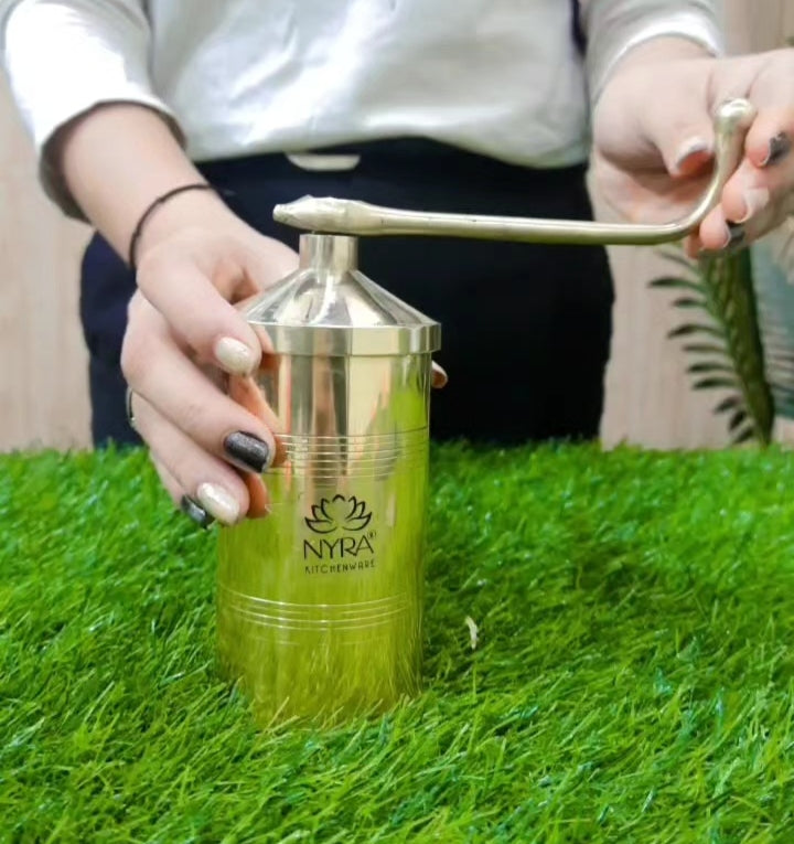 A person using a manual metal coffee grinder with a long handle, standing on artificial green grass. The brand name 'NYRA KITCHENWARE' is printed on the side of the silver-colored grinder.