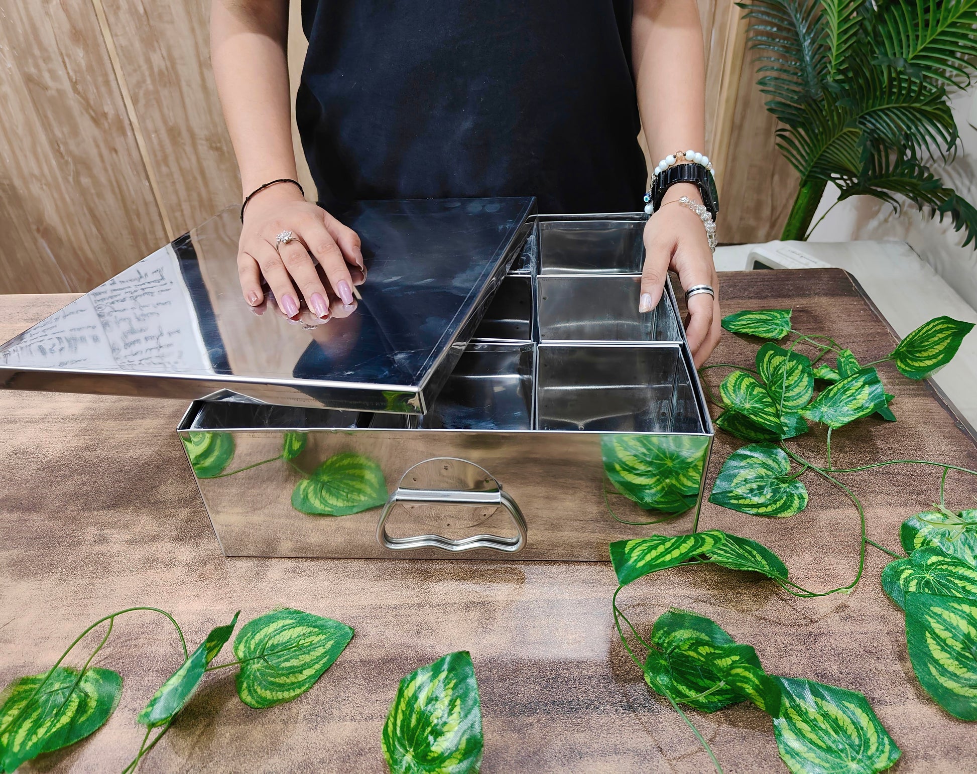 Person using a metal tray with compartments on a wooden surface with green leaves.