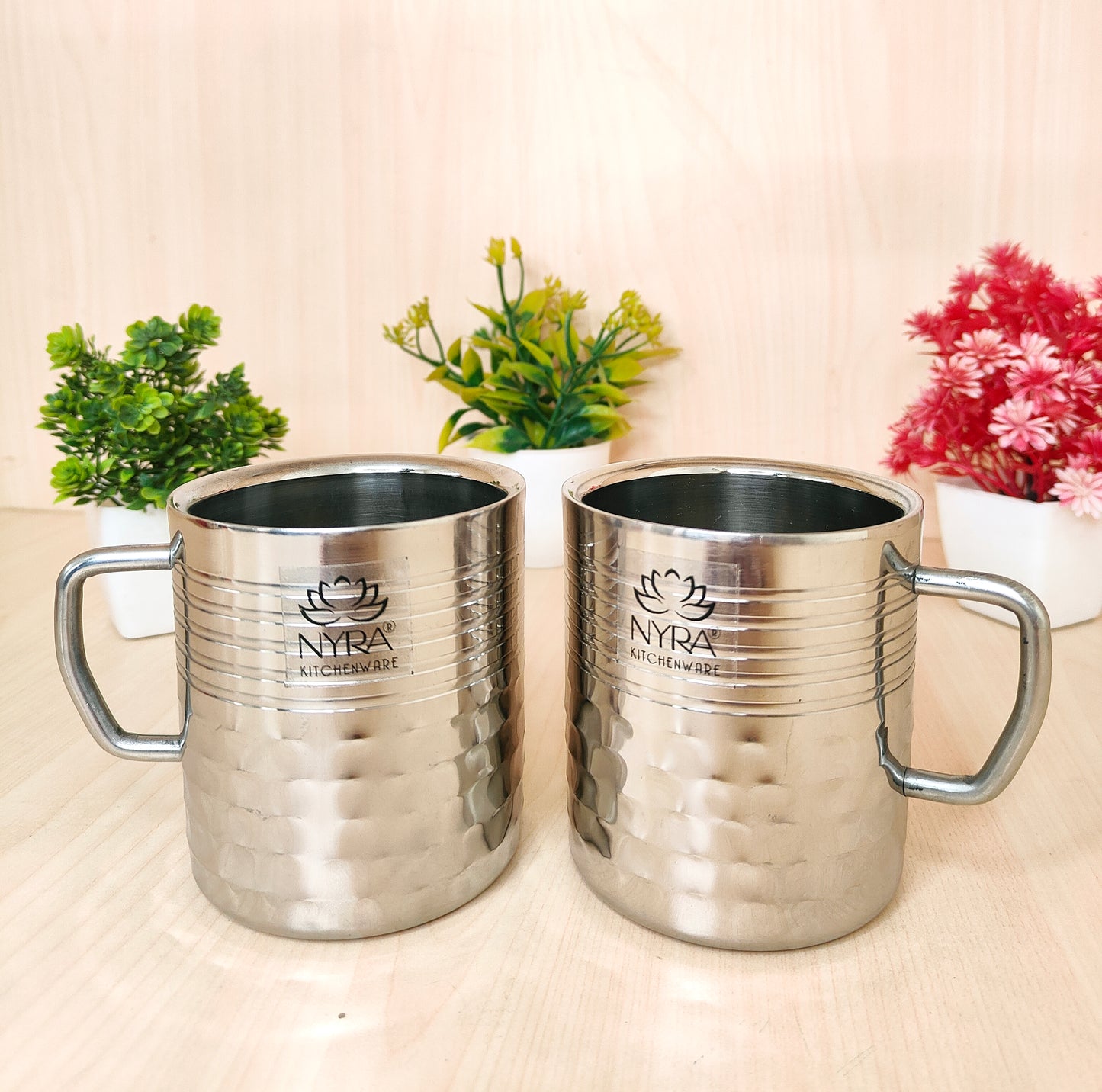 Two silver mugs with 'NYRA' branding on a light surface with plants in the background.