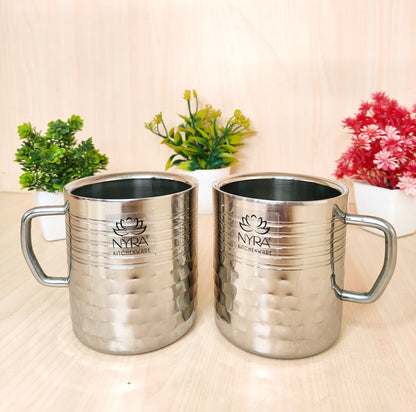 Two silver mugs with 'NYRA' branding on a light surface with plants in the background.