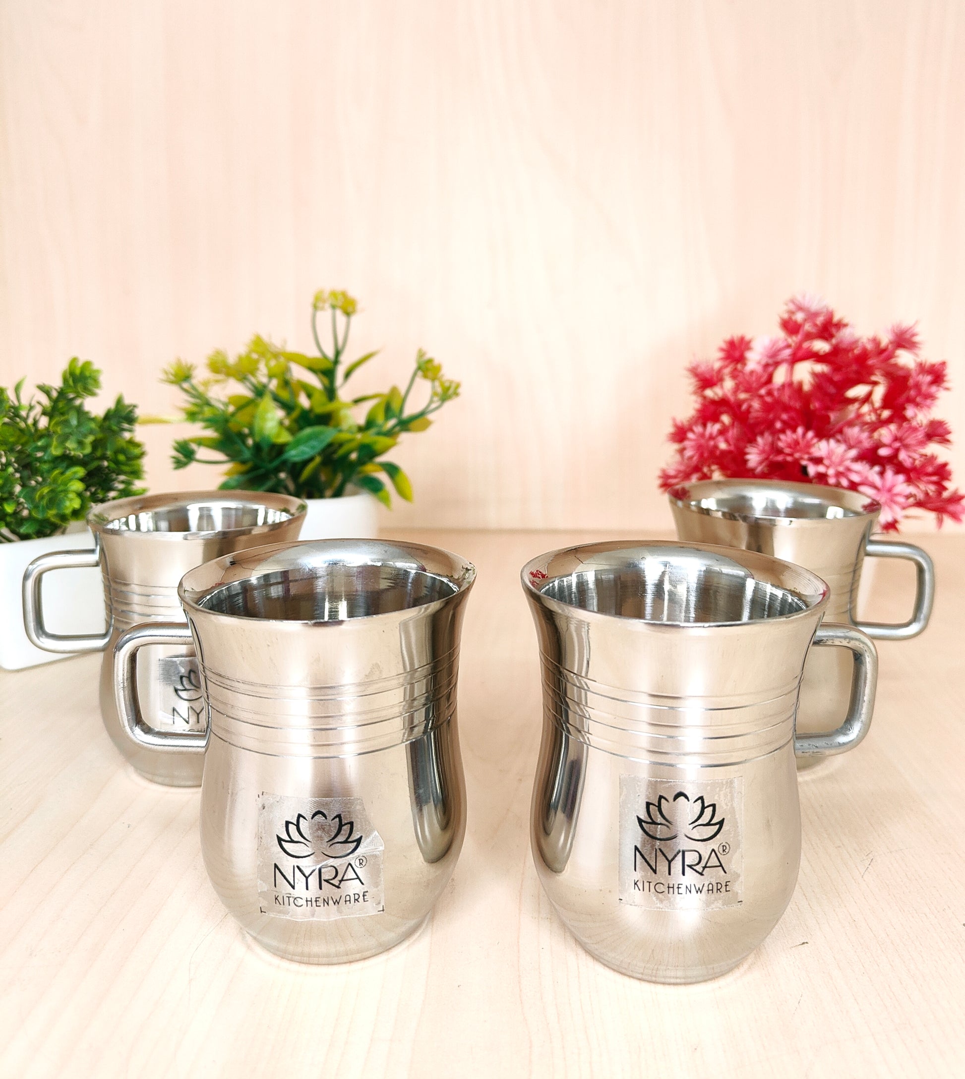 Set of silver mugs with 'Nyra' branding on a light background with plants.