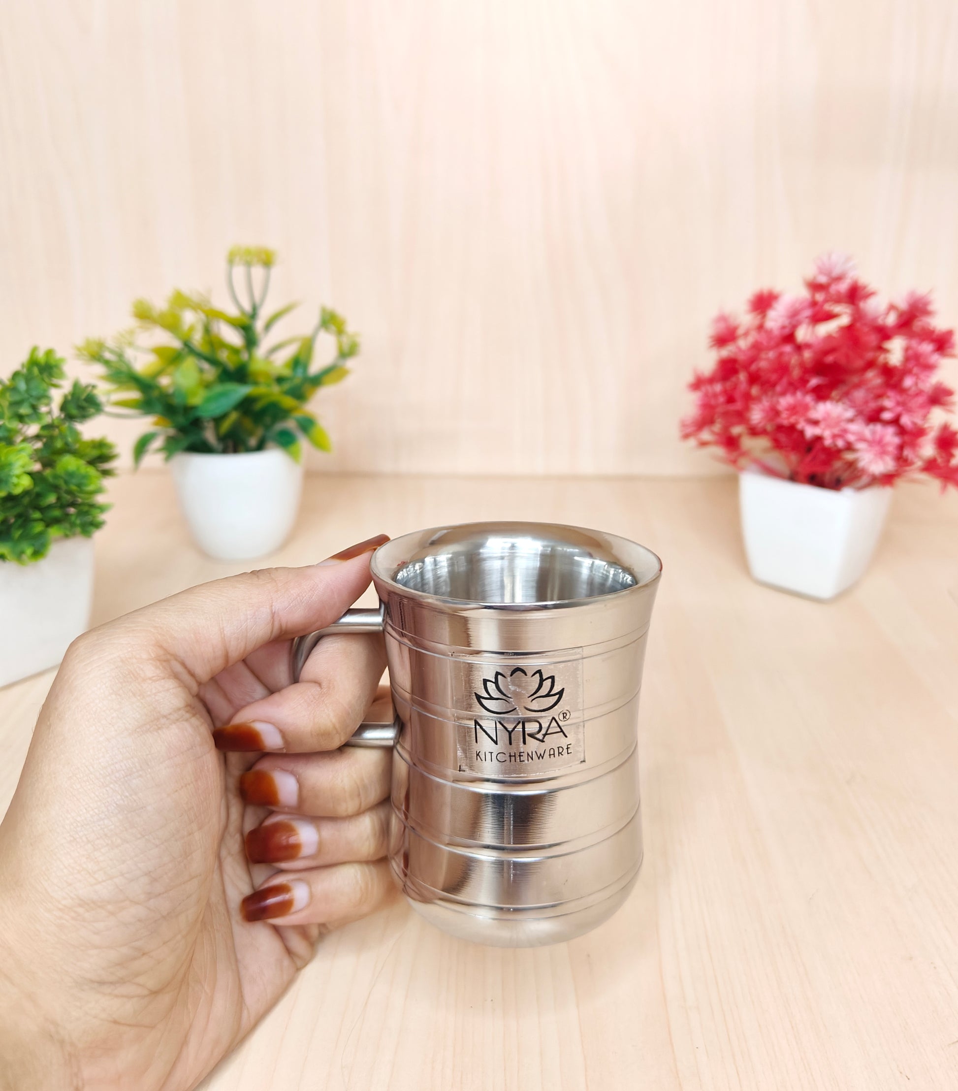 Hand holding a silver cup with 'Nyra' branding against a light background with plants.