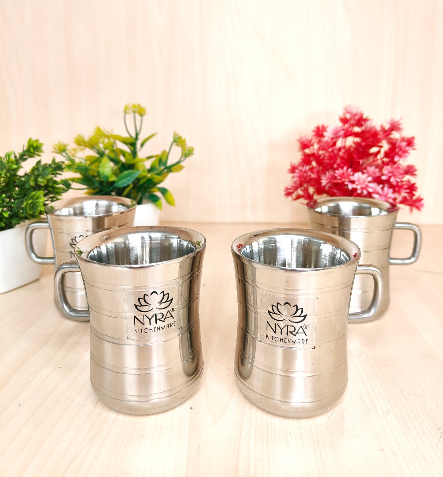 Set of silver mugs with 'Nyra' branding on a light surface with plants in the background