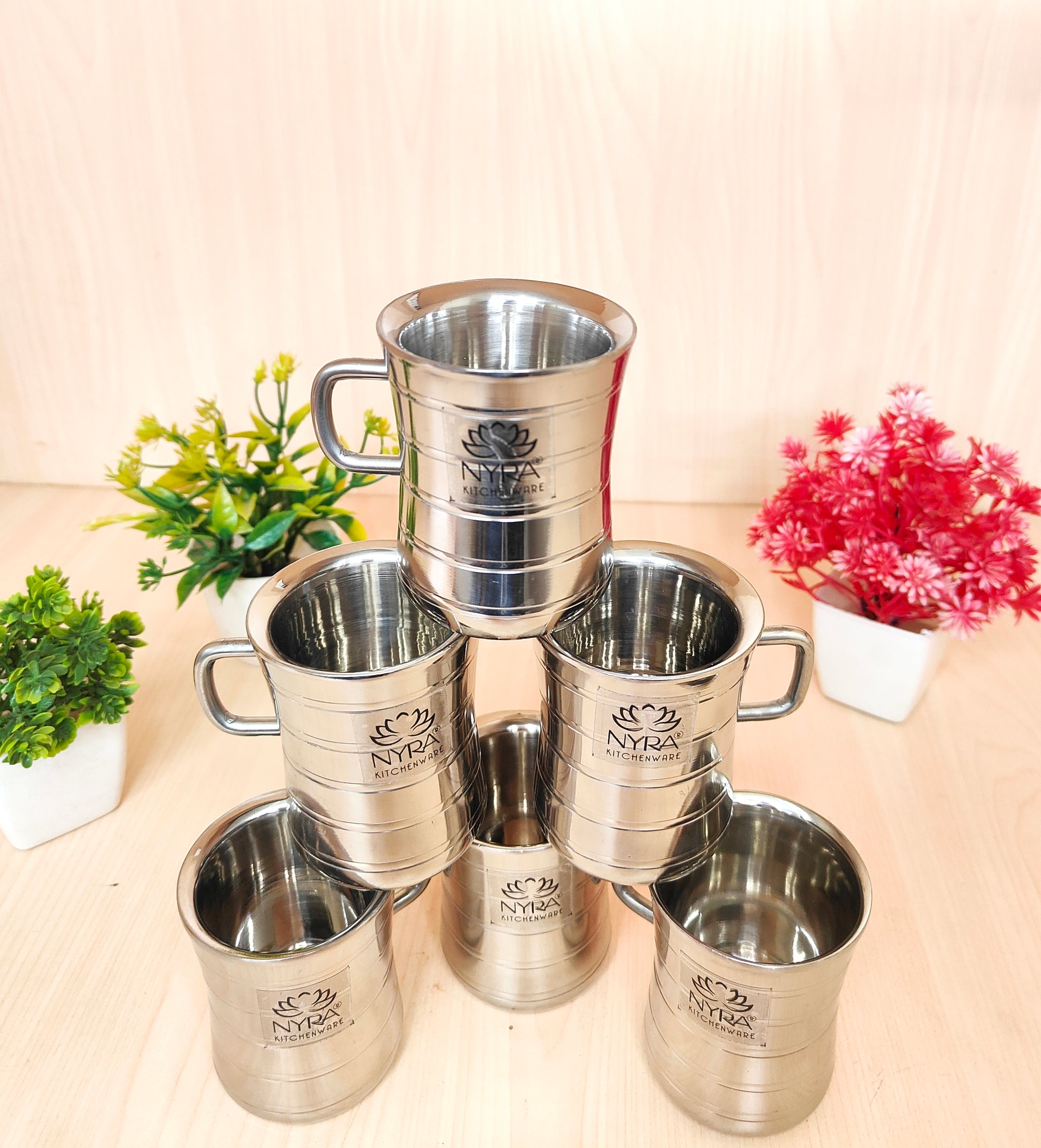 Set of silver cups with 'Nyra' branding on a light surface with plants in the background