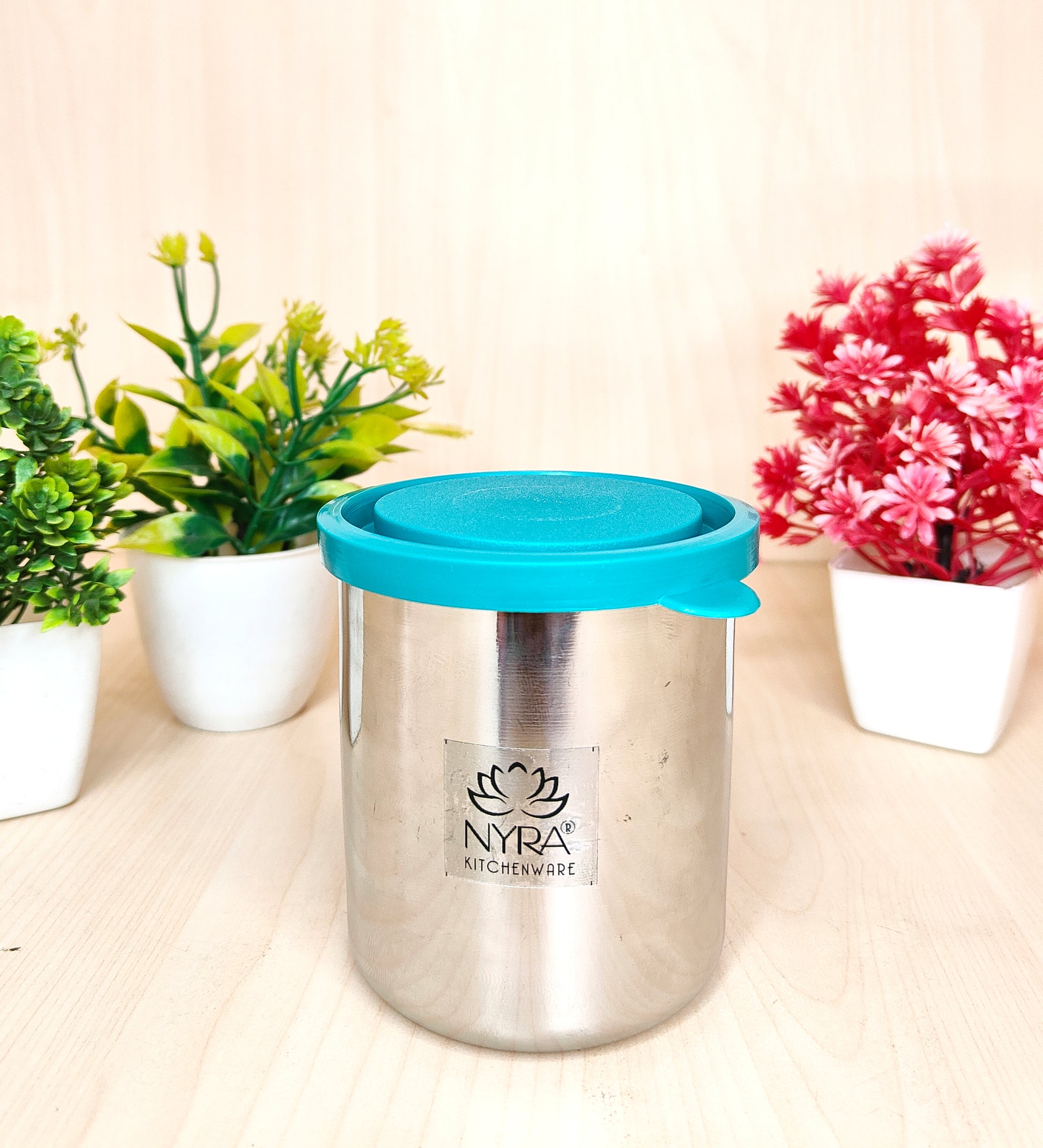 Stainless steel container with a blue lid on a light surface with plants in the background