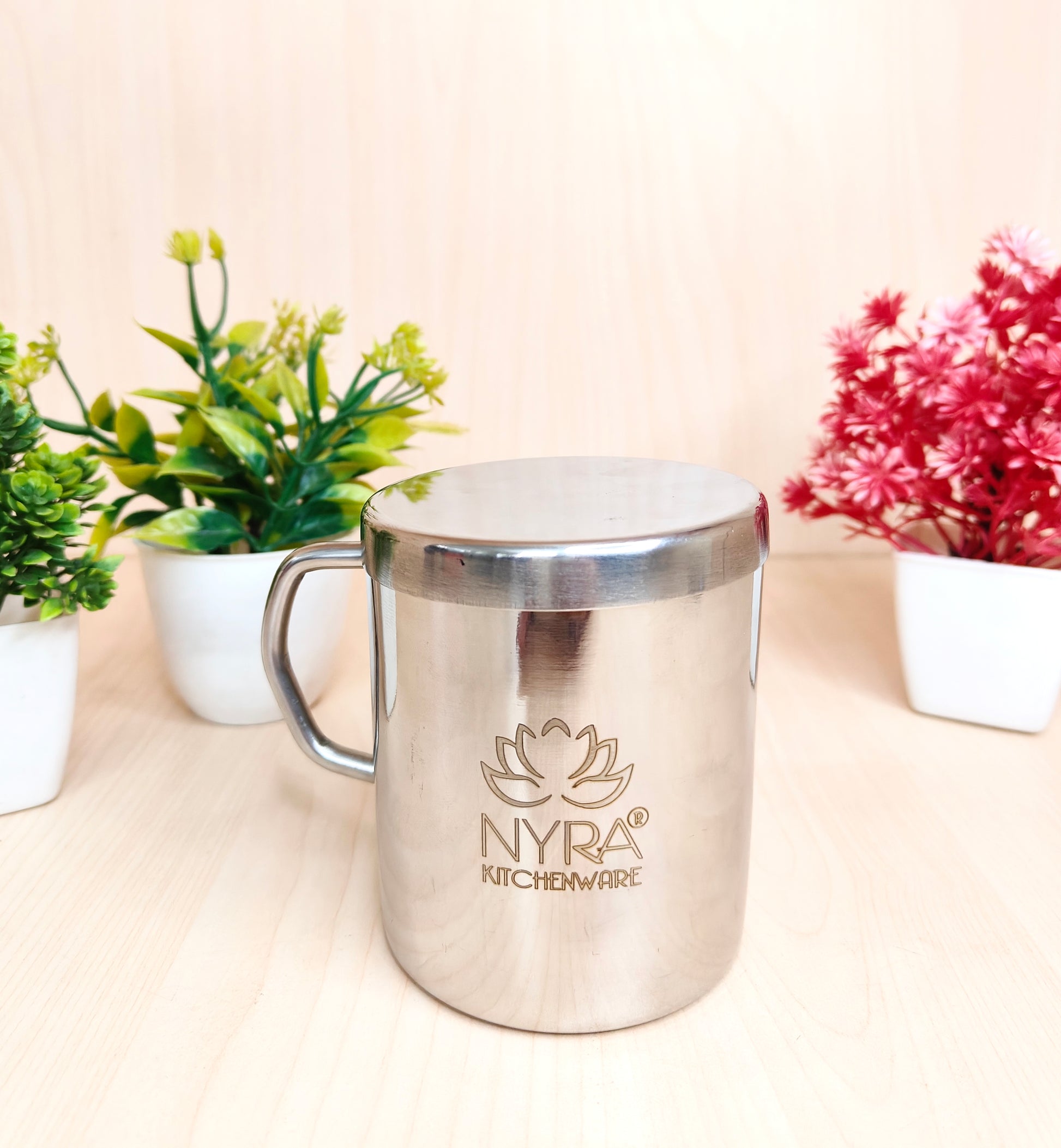 Stainless steel mug with 'Nyra Kitchenware' logo on a light surface with plants in the background