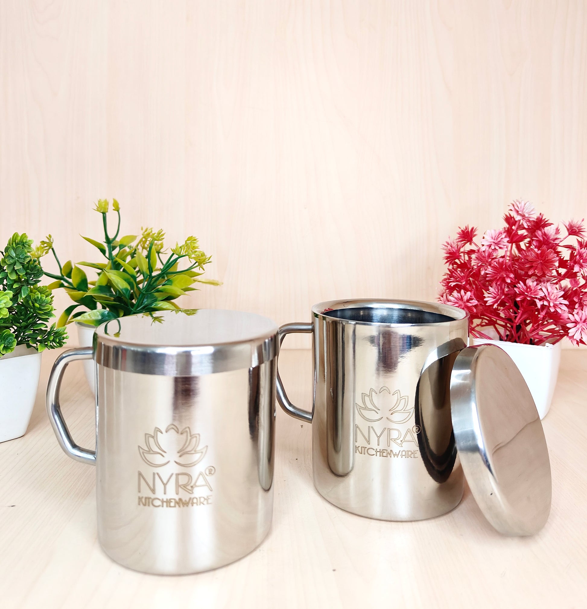 Two silver mugs with 'NYRA' branding on a light surface with plants in the background.