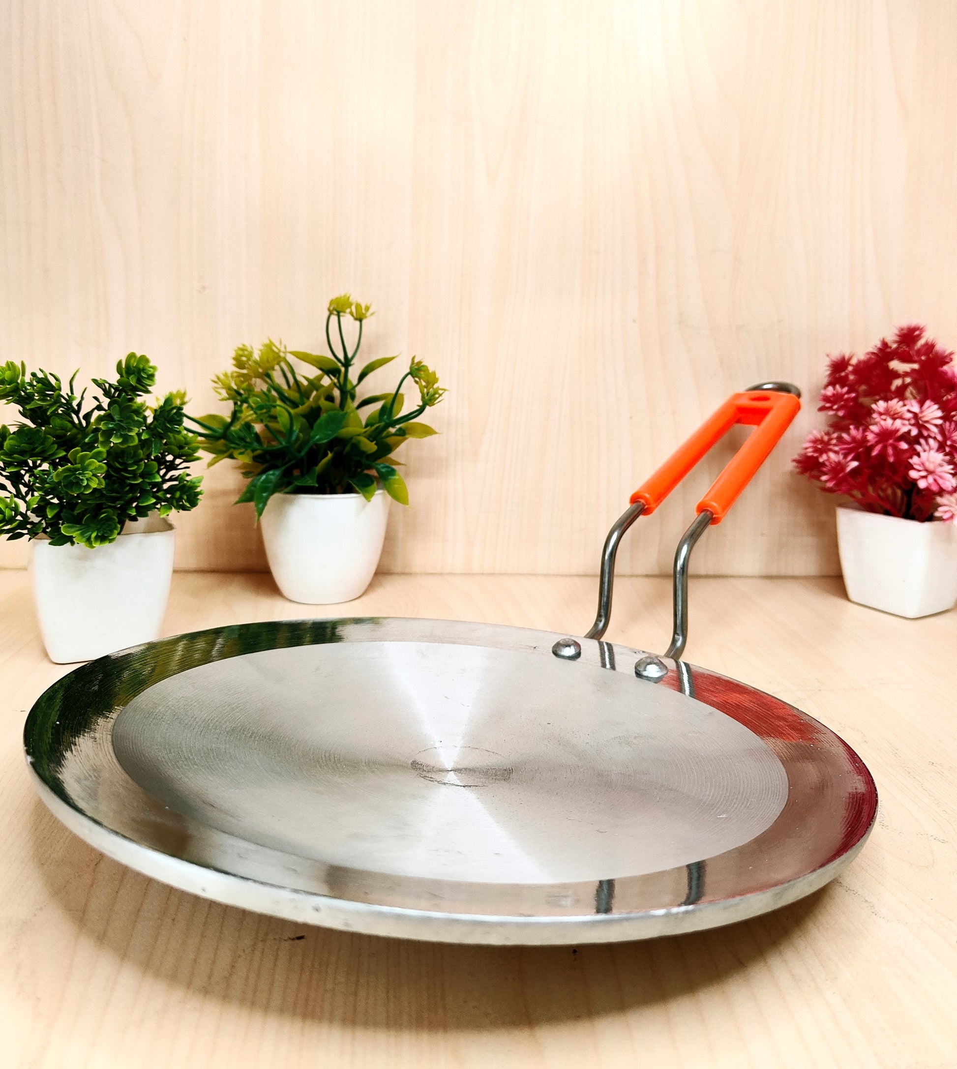 Stainless steel frying pan with orange handle on a wooden surface with decorative plants.