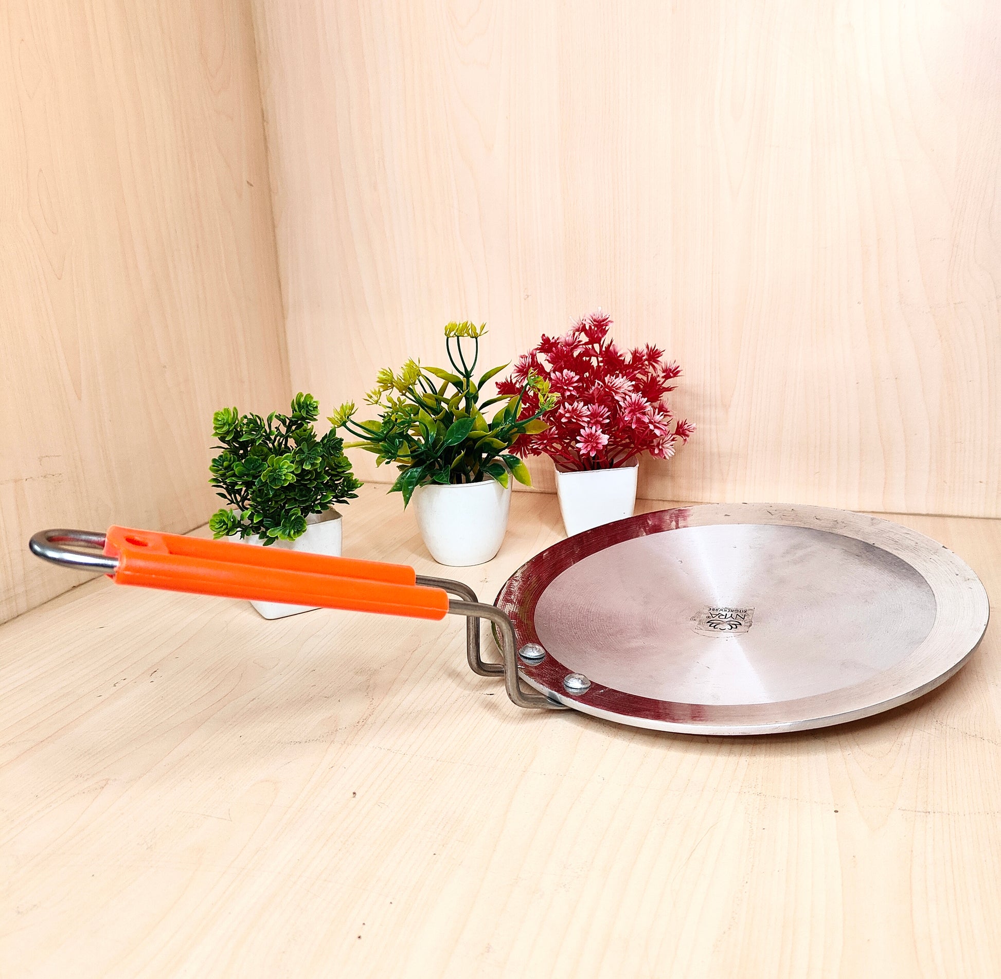 Metal frying pan with orange handle on a wooden surface with plants in the background
