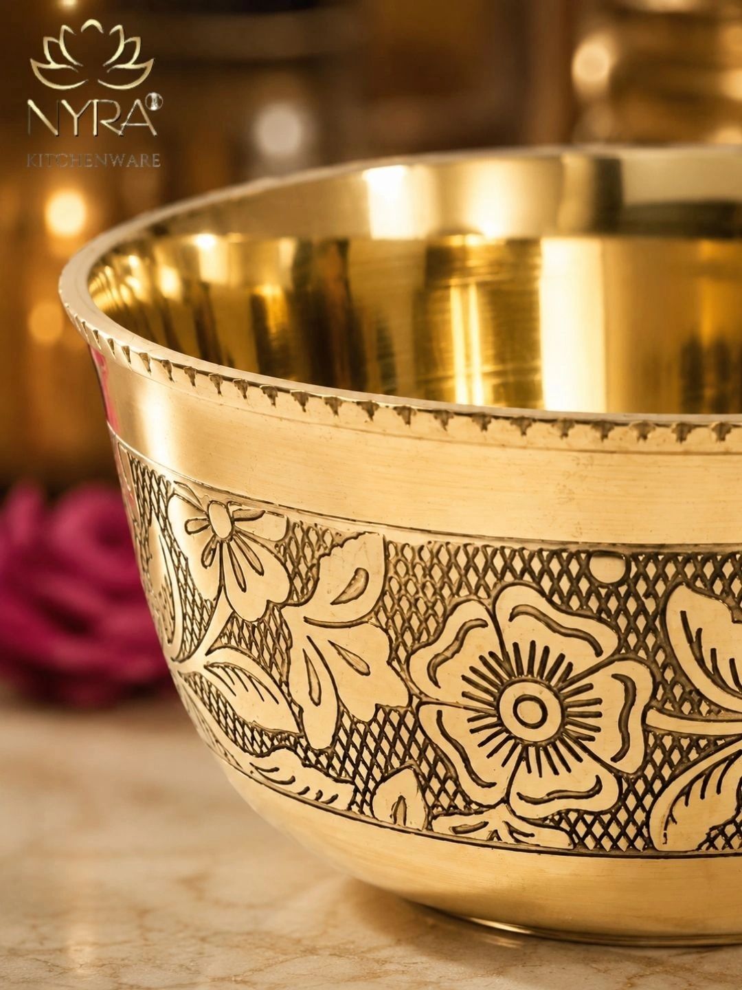 A close-up of a polished brass bowl with intricate floral engravings on the exterior, set against a warm, softly blurred background. The NYRA brassware logo is visible in the top-left corner.