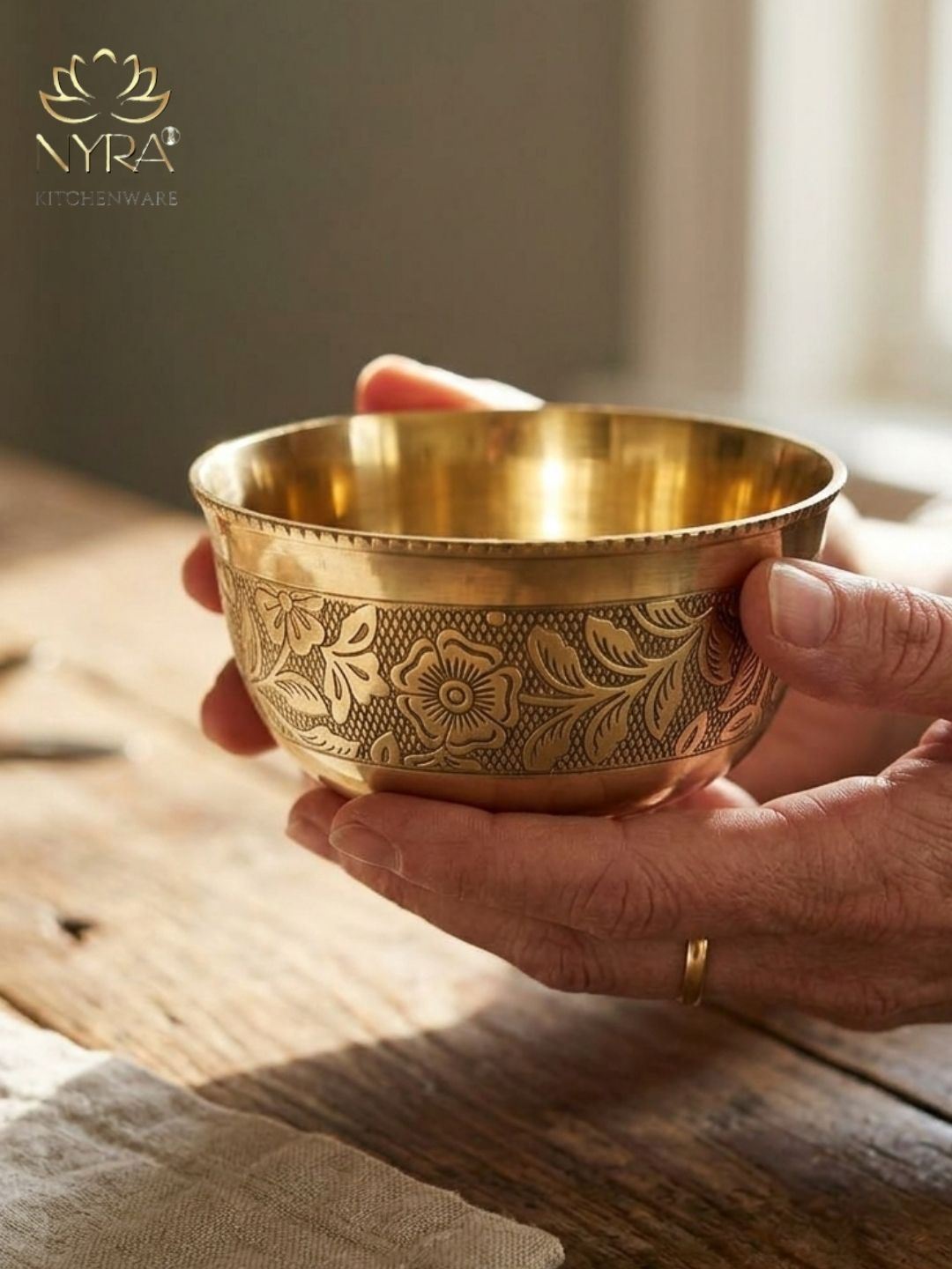 A hand holds a small brass bowl with floral engravings, set against a rustic wooden table background with a soft-focus indoor setting. The NYRA brand logo is visible in the top left corner.