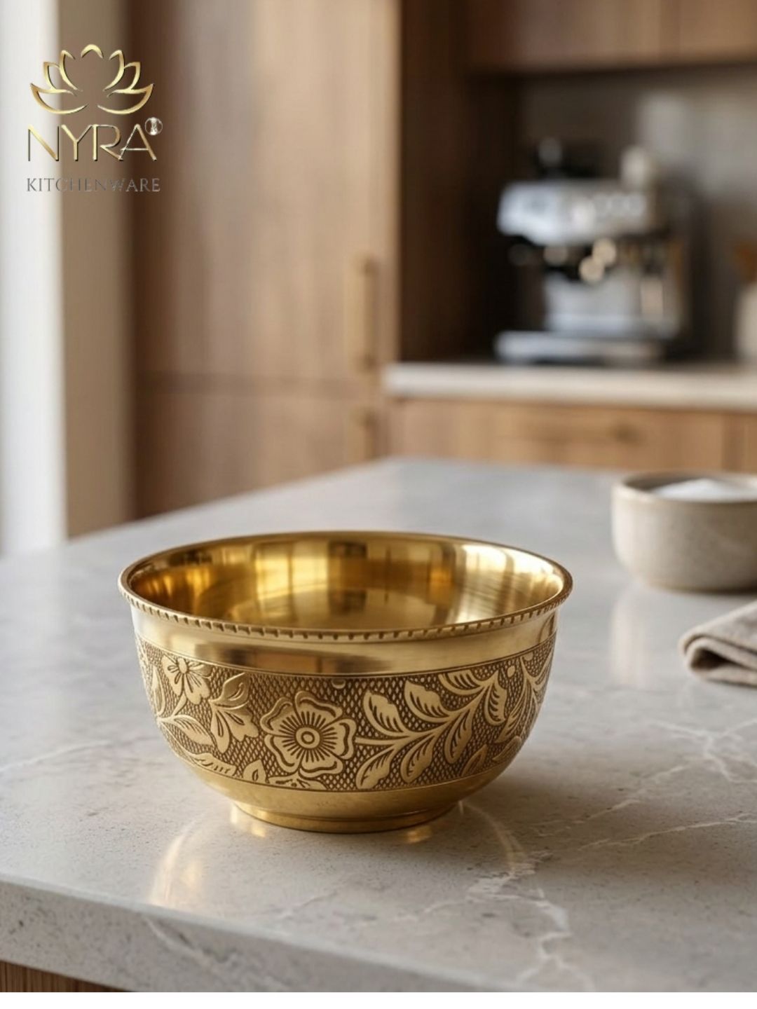 A gold-toned metal bowl with an embossed floral pattern, set on a light marble countertop in a blurred kitchen setting. The NYRA Kitchenware logo is visible in the top left corner.