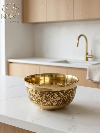 A decorative brass bowl with floral engravings sits on a white marble kitchen island, with a blurred modern kitchen background featuring light wood cabinets and a gold faucet. The NYRA KITCHENWART logo is visible in the top left corner.