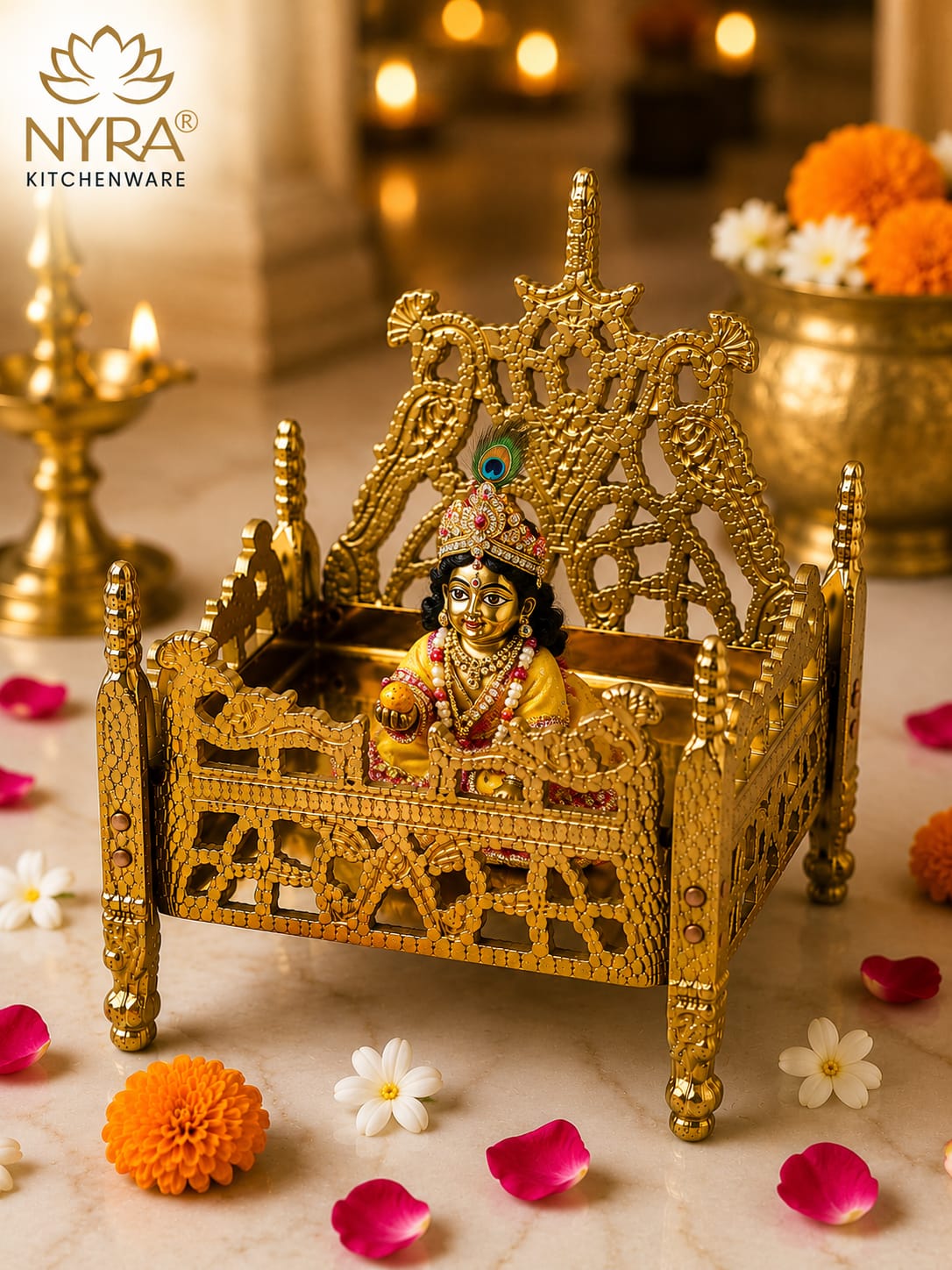 A golden ornate miniature bed cradle figurine holding a small Lord Krishna idol, styled on a marble surface with marigold flowers, rose petals, and a diya in the background. The NYRA Kitchenware logo is visible in the top left corner.