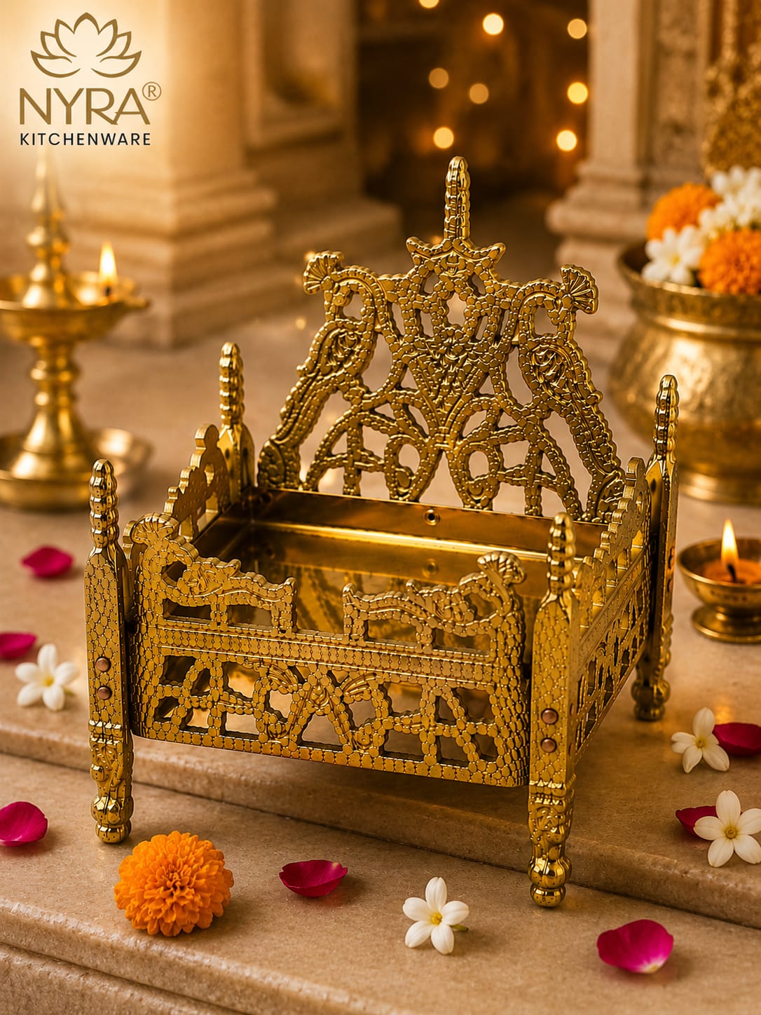 An ornate gold-toned brass miniature temple palu or decorative vessel with intricate cutout details, displayed on a stone surface with marigold petals, flowers, and lit oil lamps in a warm, festive setting. The NYRA Kitchenware logo is visible in the top left corner.